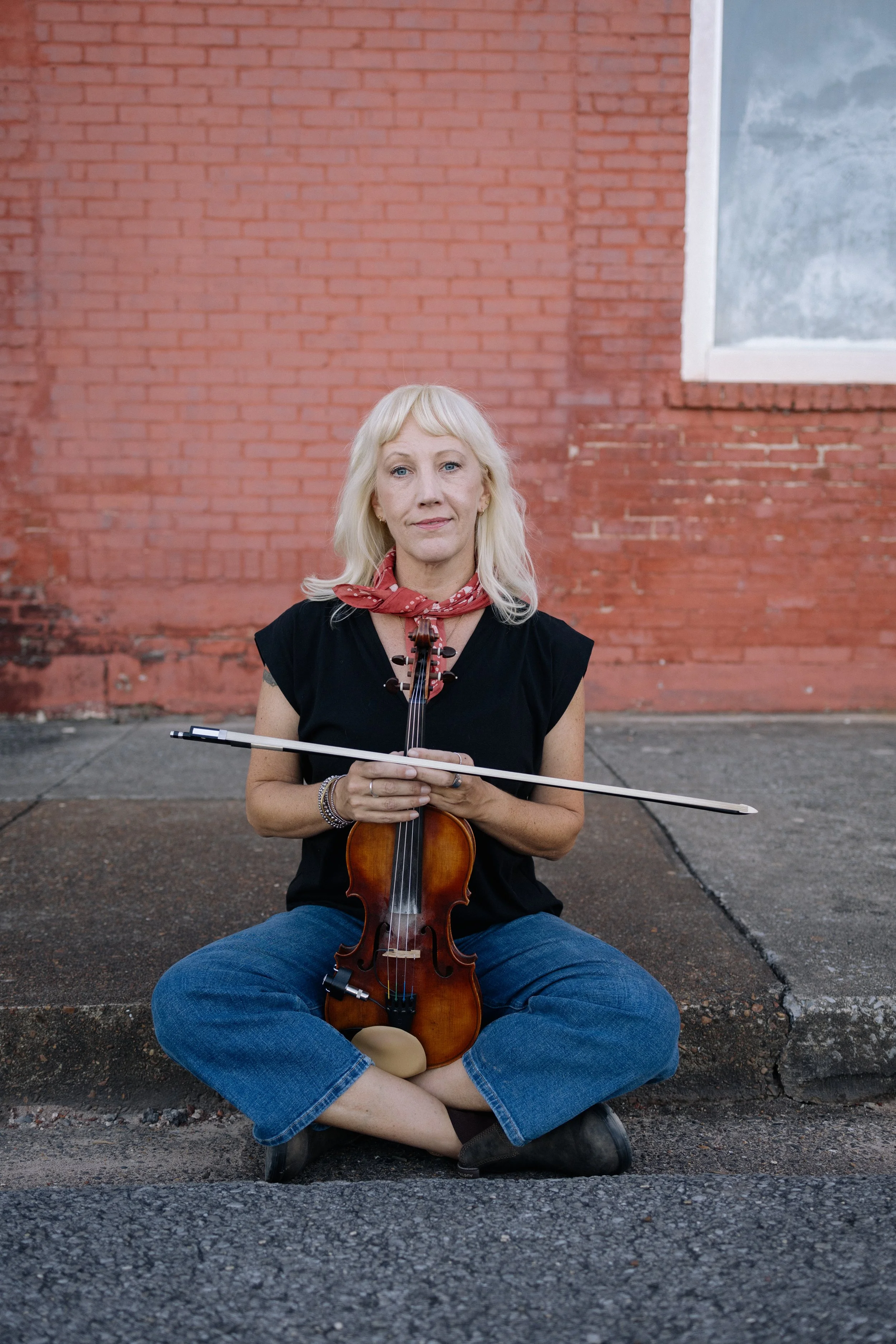A woman with blonde hair and a red bandana around her neck is sitting cross-legged on the pavement outside, holding a violin and a bow, with a brick wall and a window with a snowy landscape visible in the background.