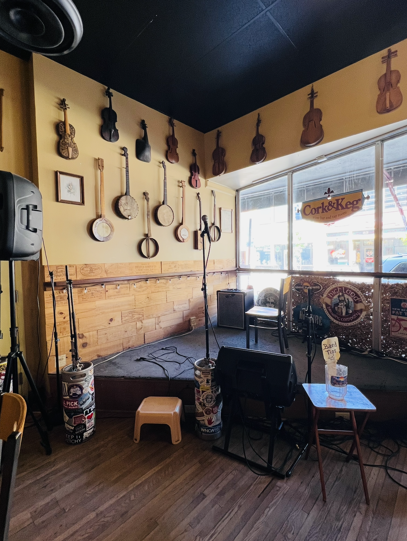 Interior view of a small music venue or bar, with a yellow wall decorated with various stringed instruments such as violins and banjos. A stage setup with microphones, speakers, and stools is visible. Outside, through large windows, a sign for the es