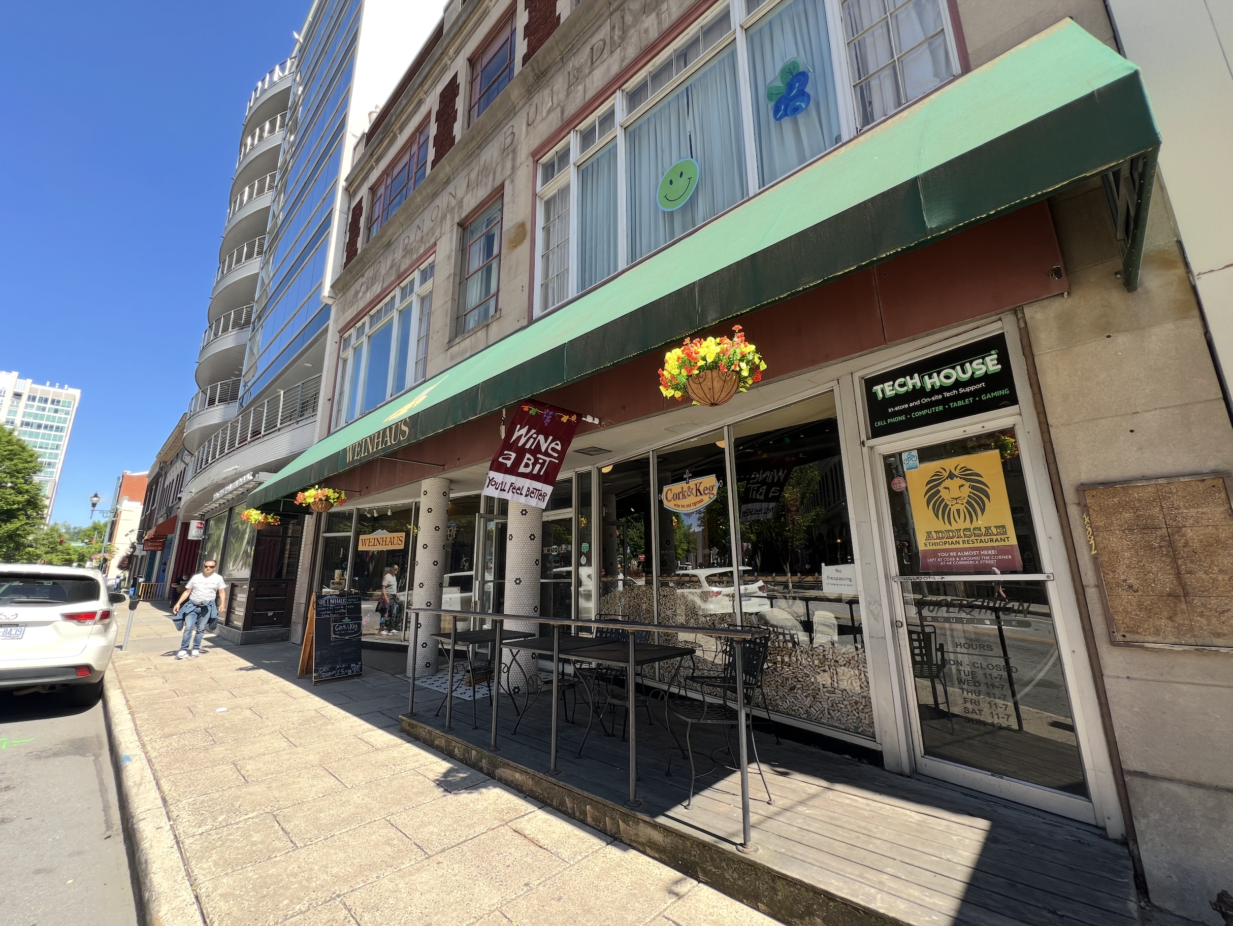 Storefront with green awning and outdoor seating, with signage for Wein Haus and Tech House, and decorations including hanging flower baskets and smiley face and balloon window decals.