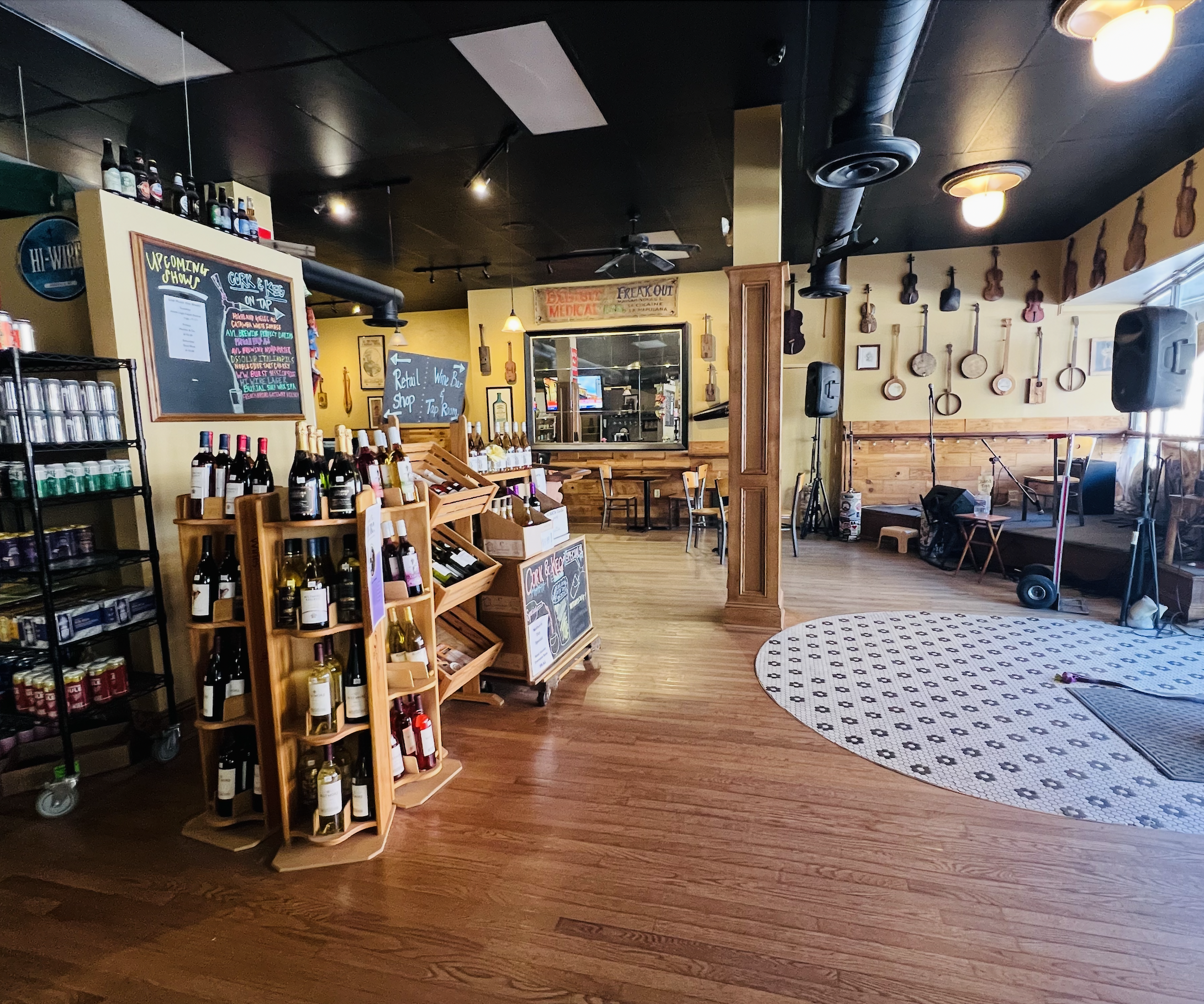 A cozy indoor space resembling a bar or cafe with wooden flooring, wine bottles on display, and musical instruments hanging on the wall. There are tables, chairs, and stage lighting, suggesting a performance area.