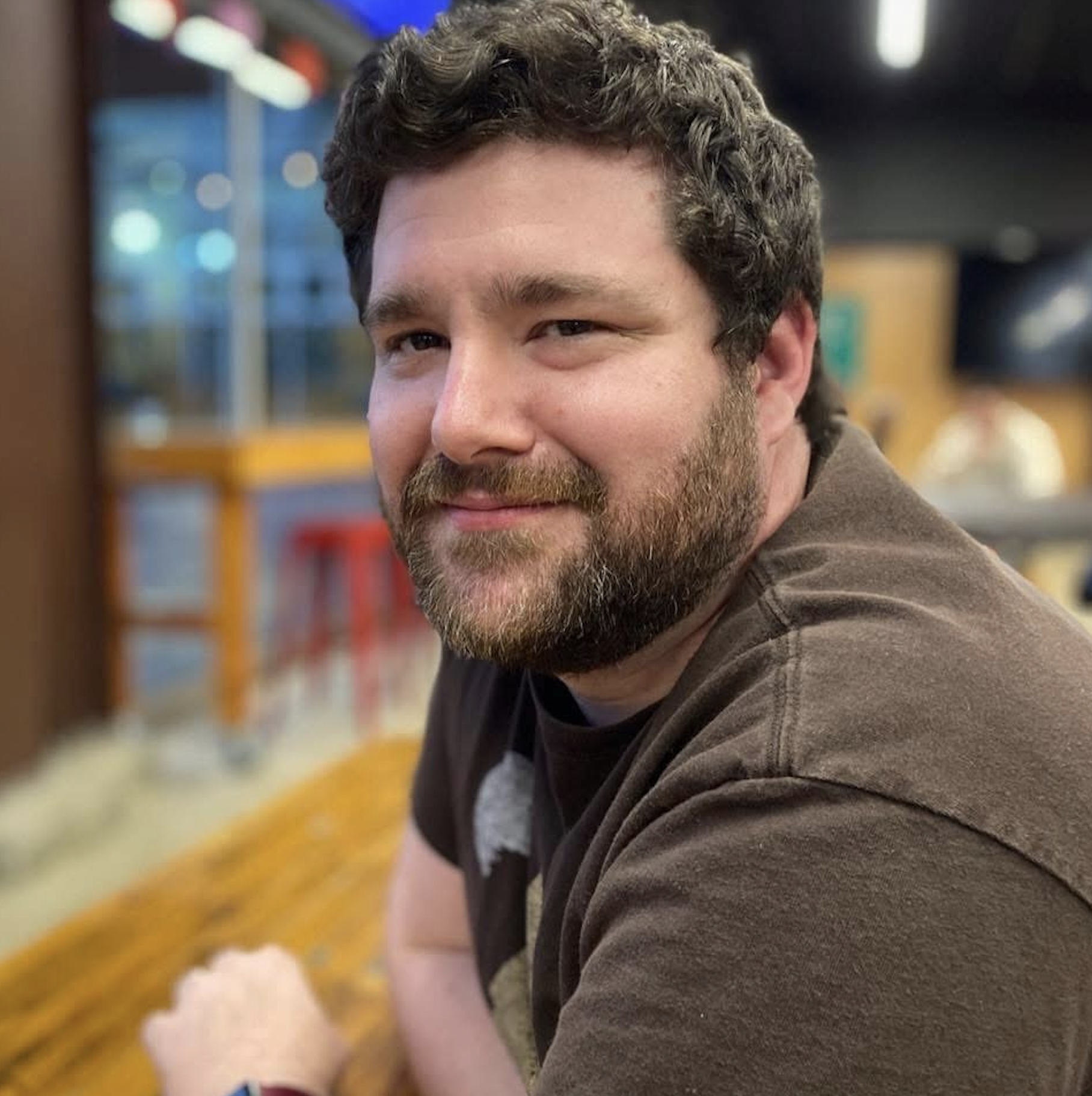 A man with brown curly hair and a beard, smiling and looking to the side, in a casual setting with colorful blurred background.