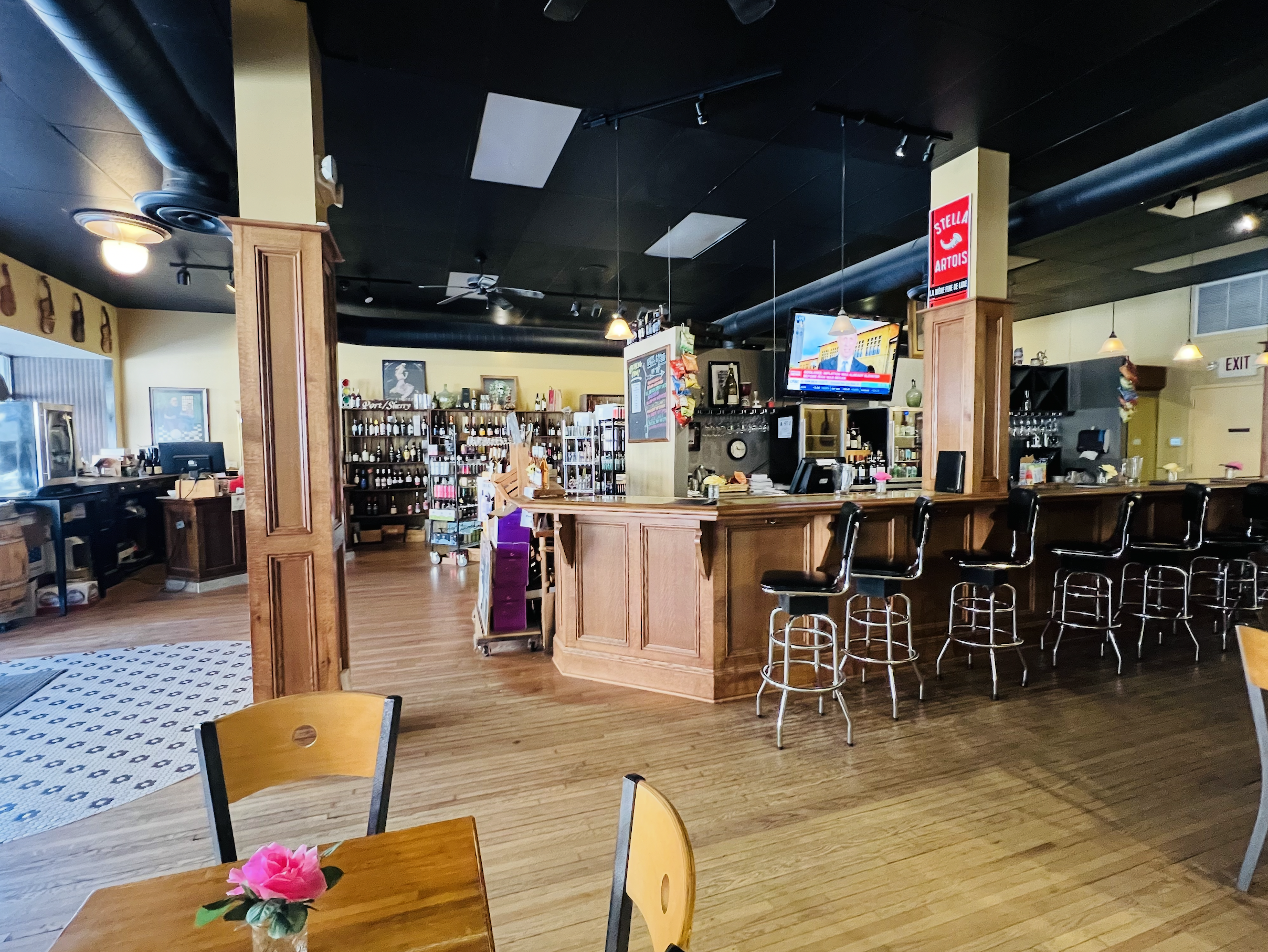Interior of a bar or restaurant with wooden counters, black bar stools, a TV displaying a news broadcast, shelves with bottles, and decorative elements.
