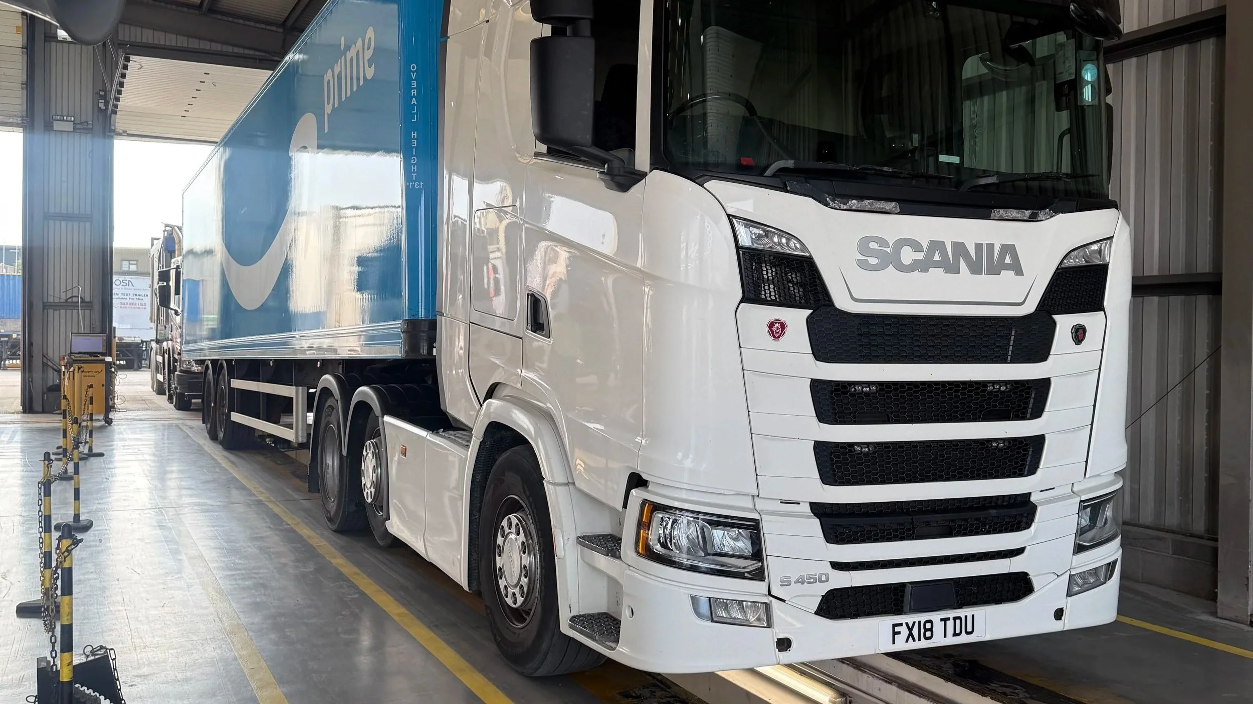 A white Scania truck parked inside a warehouse or garage, connected to a large blue delivery trailer with Amazon Prime branding.