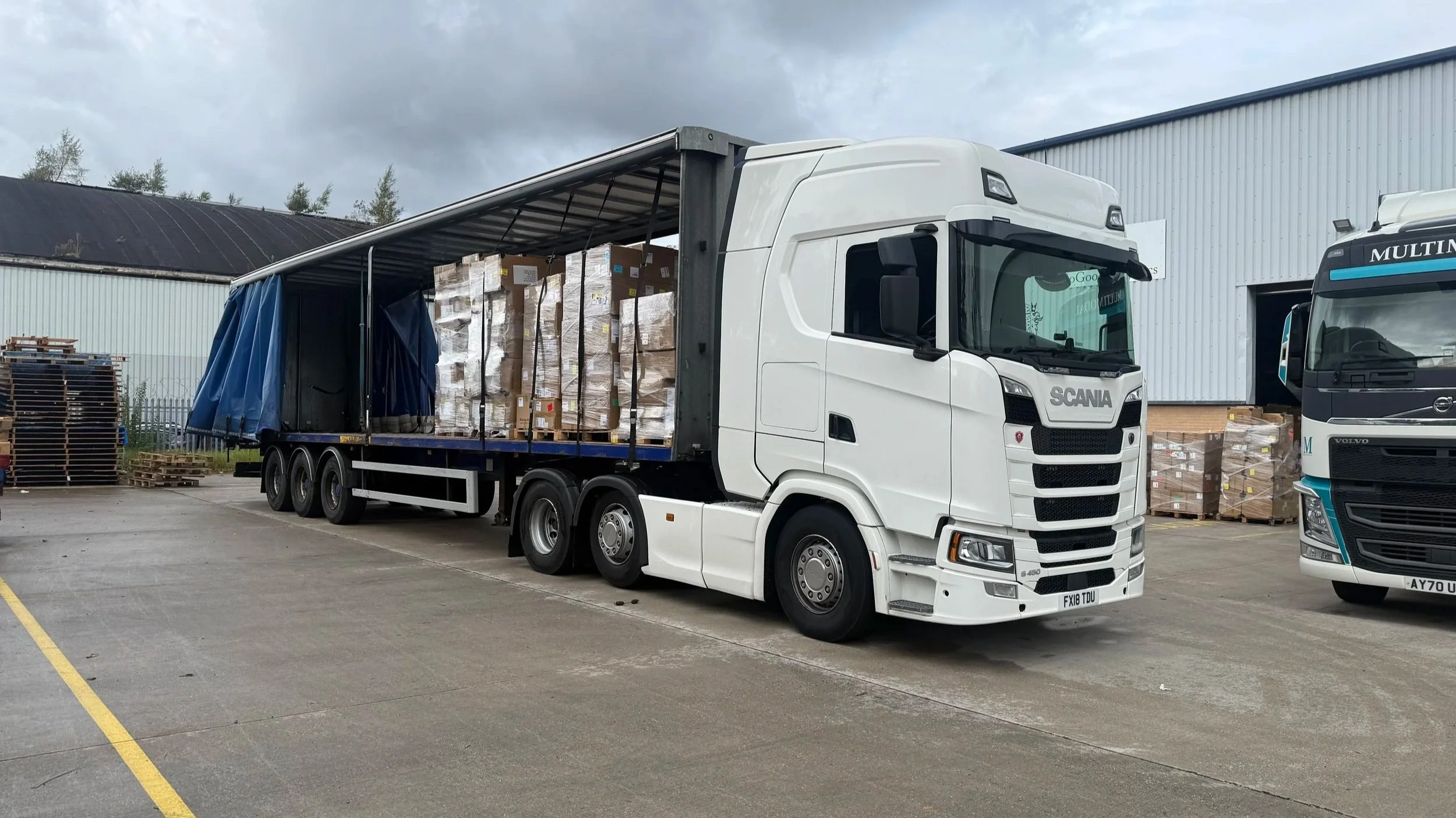 A white semi-truck with an open loading area filled with boxes, parked outdoors near a warehouse, with another truck and stacks of pallets nearby.