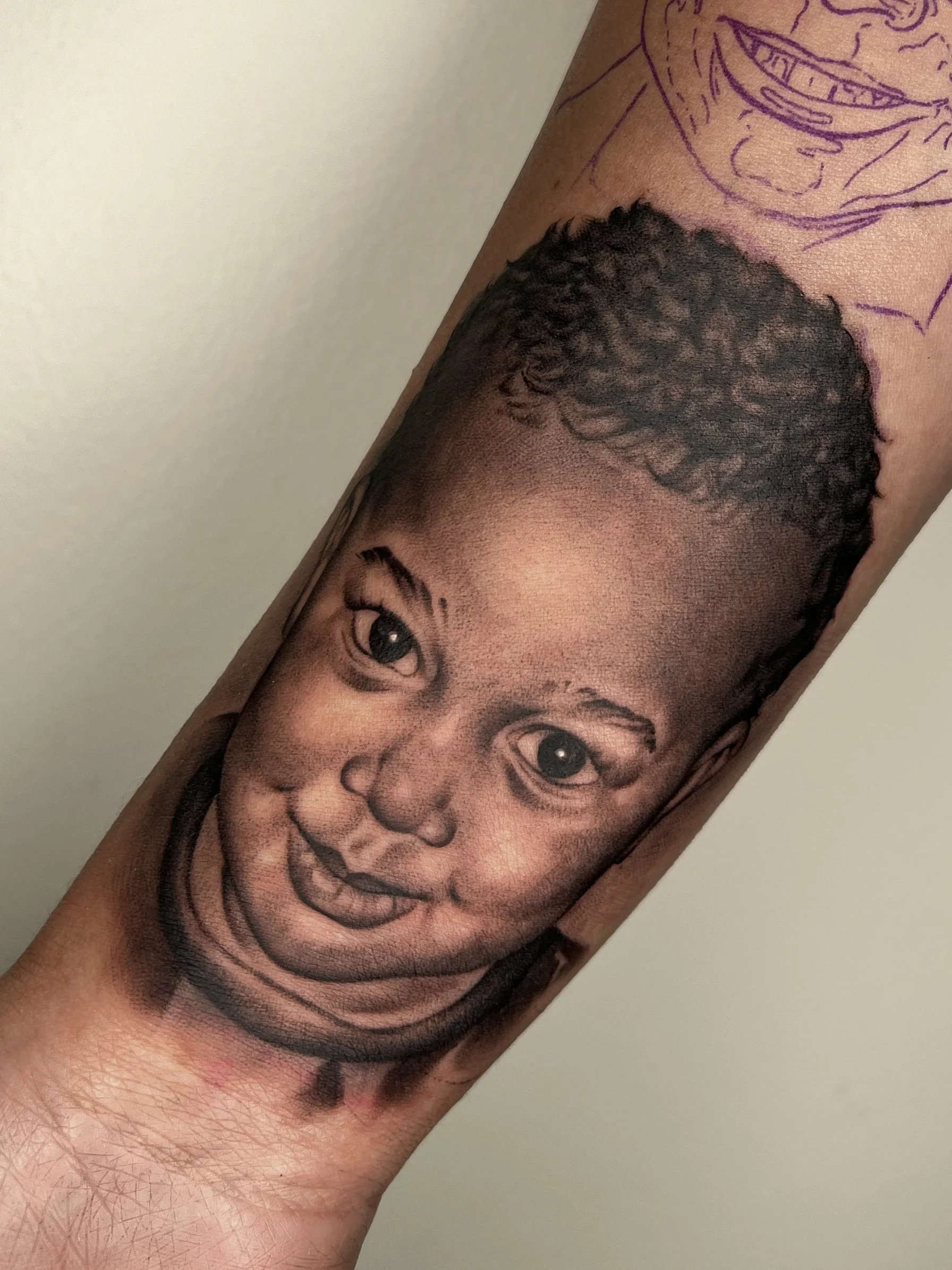 A tattoo of a realistic child's face on a person's arm, with a partial drawing of a face outline above it.