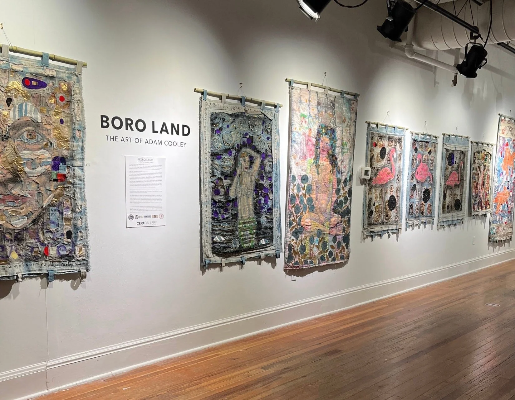 A gallery wall displays multiple fabric art pieces by Adam Cooley, titled 'Boro Land,' featuring intricate textile designs, embroidered patterns, and vibrant colors, with informational signage on the wall.