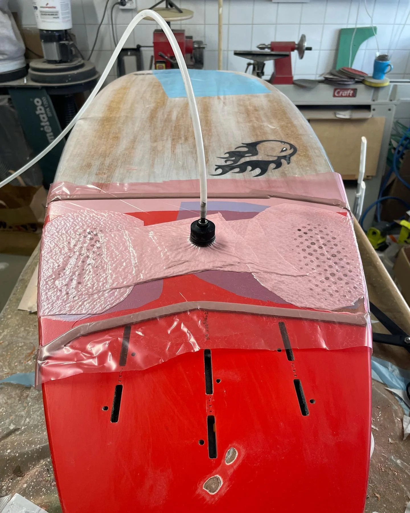 En surfbräda under reparation, placerad på en röd arbetsbänk i ett verkstadsskriv. Surfbrädan är delvis täckt med plastfilm och har ett bilder av en svart drake på toppen. En svart slang är ansluten till brädan. Verktyg och maskiner syns i bakgrunden.