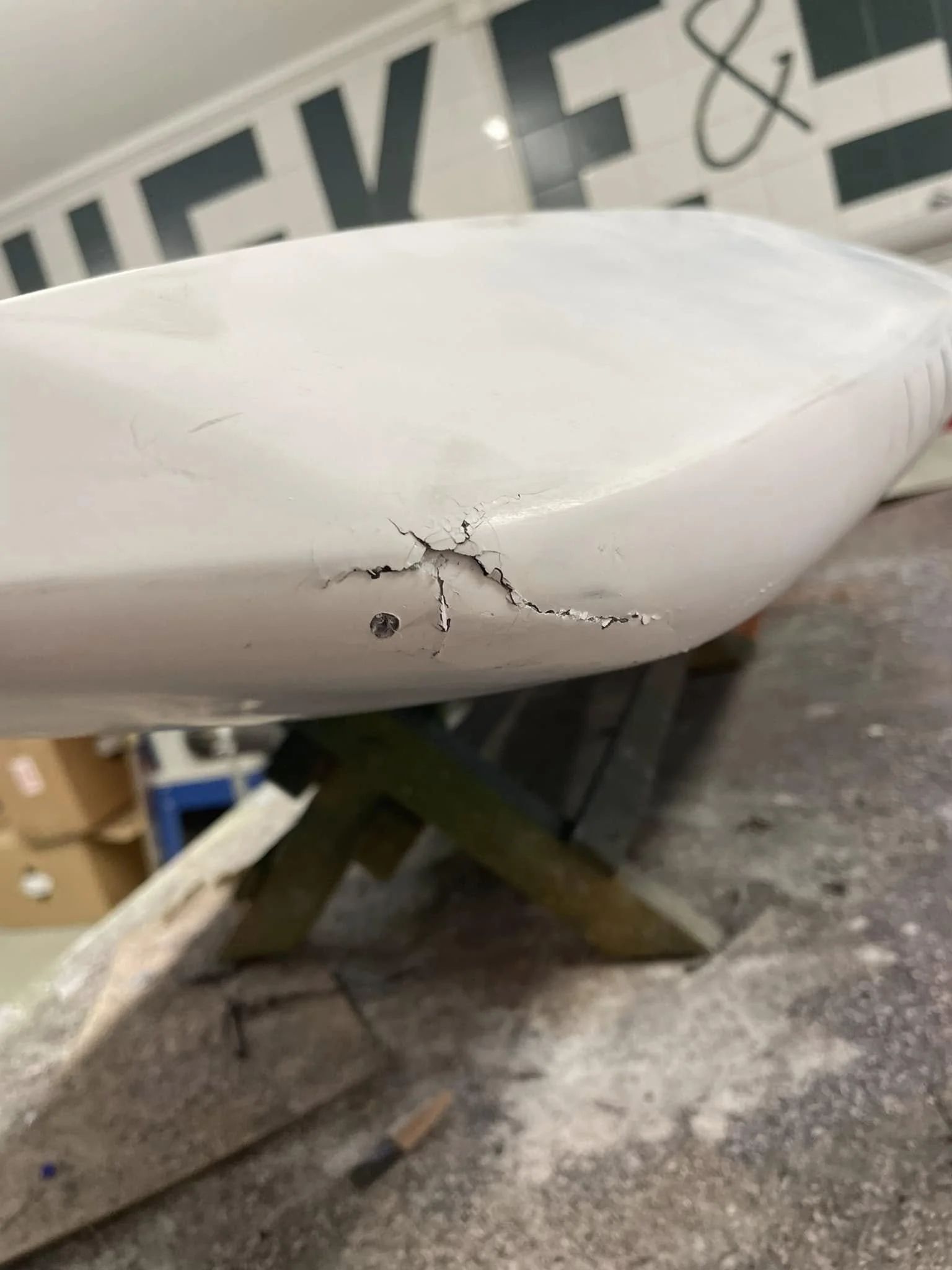 En vit yta med ett sprucket och skadat område, antagligen av plast eller keramik, placerad på ett träbord. Bakom finns en skog av svarta och vita rutor, troligen del av en väggdekoration eller mural.