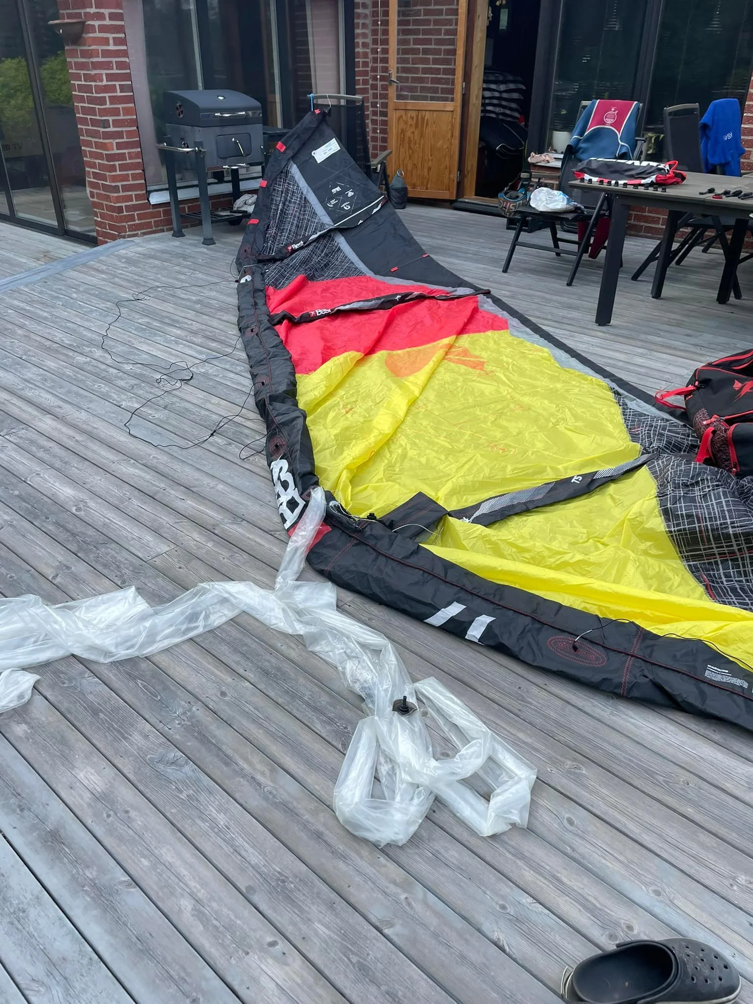 Utrustning för kite surfing  inklusive ett rödmålat, gult och svart kite, bladder, och annat tillbehör på en träterrass utanför ett hus.