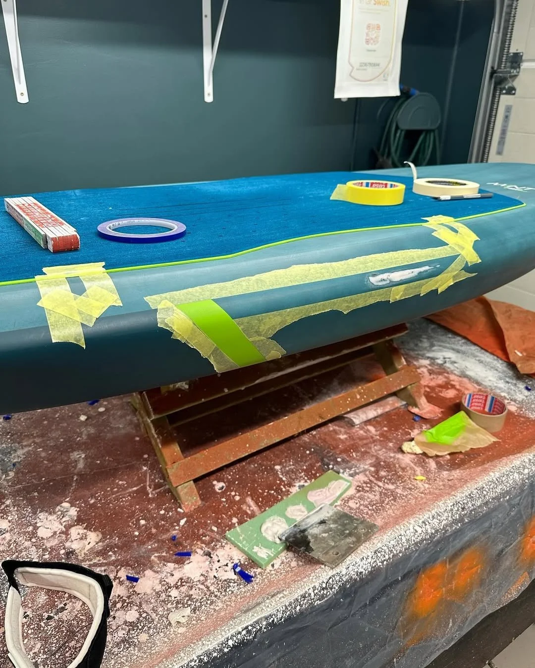 En vindsurfbräda som är i färd med att repareras, med tejp och material för reparation på och runt surfbrädan, placerad på en arbetsbänk.