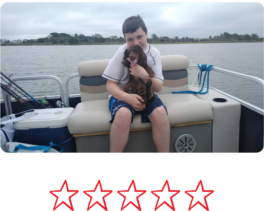 A boy sitting on a boat seat holding a brown puppy, with water and trees in the background.