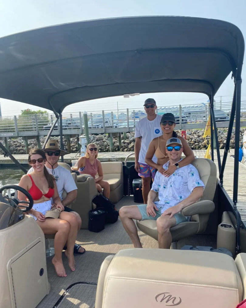 Six people enjoying a day on a boat at a marina, some sitting and some standing, smiling at the camera. The boat has a canopy overhead and the background shows water, rocks, and a dock.