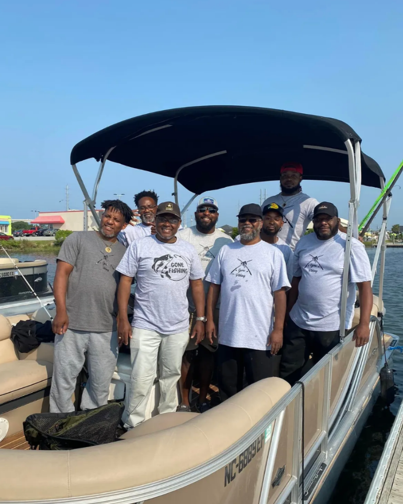 Group of nine men on a boat, some wearing grey t-shirts, one with a 'Gone Fishing' graphic, standing on a boat docked on water with a marina and buildings in the background, under a clear blue sky.