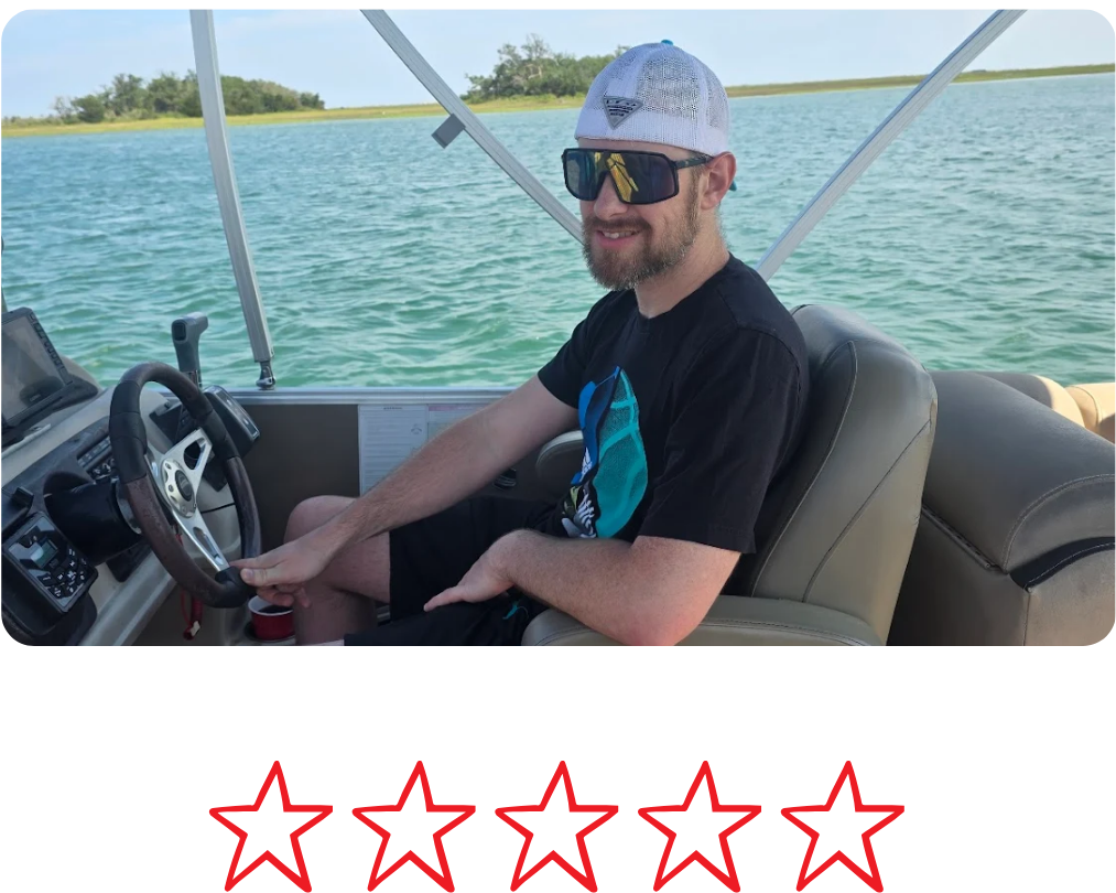 A man sitting at the wheel of a boat on the water, wearing sunglasses, a baseball cap, and a black t-shirt, with a body of water and trees in the background.
