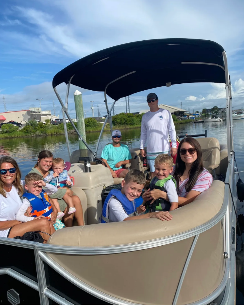 A group of children and adults on a boat in a harbor, with some children wearing life jackets, a woman in sunglasses, a man in a white long-sleeve shirt, and a sunny sky with some clouds.