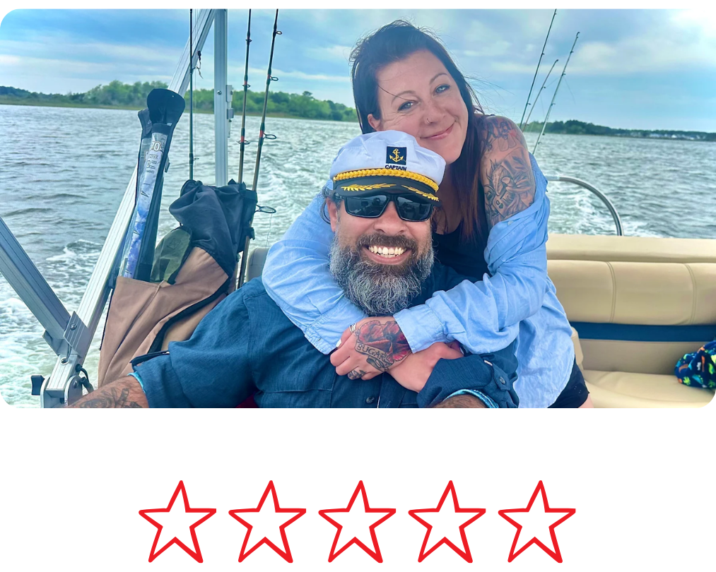A couple enjoying a boat ride on a lake, smiling and hugging. The woman has tattoos, and the man is wearing a captain's hat and sunglasses. The boat has fishing rods and a bag in the background, with water and trees visible in the distance.