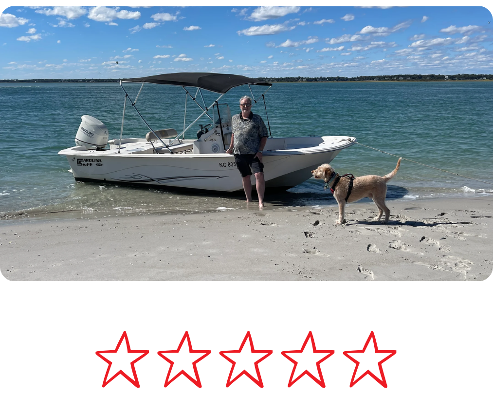 A man with sunglasses and a patterned shirt standing on the beach beside a small white boat with a dog on a leash nearby, clear blue water and partly cloudy sky in the background.