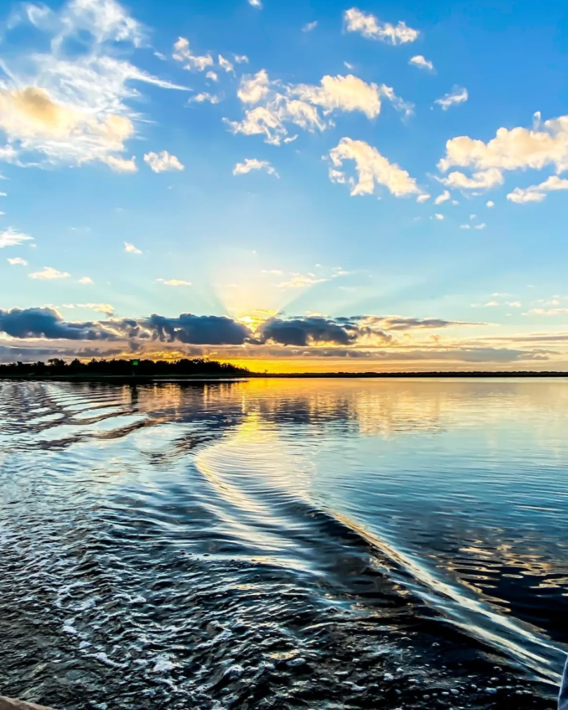 Sunset over a large body of water with a partly cloudy sky, and a boat's wake rippling behind.