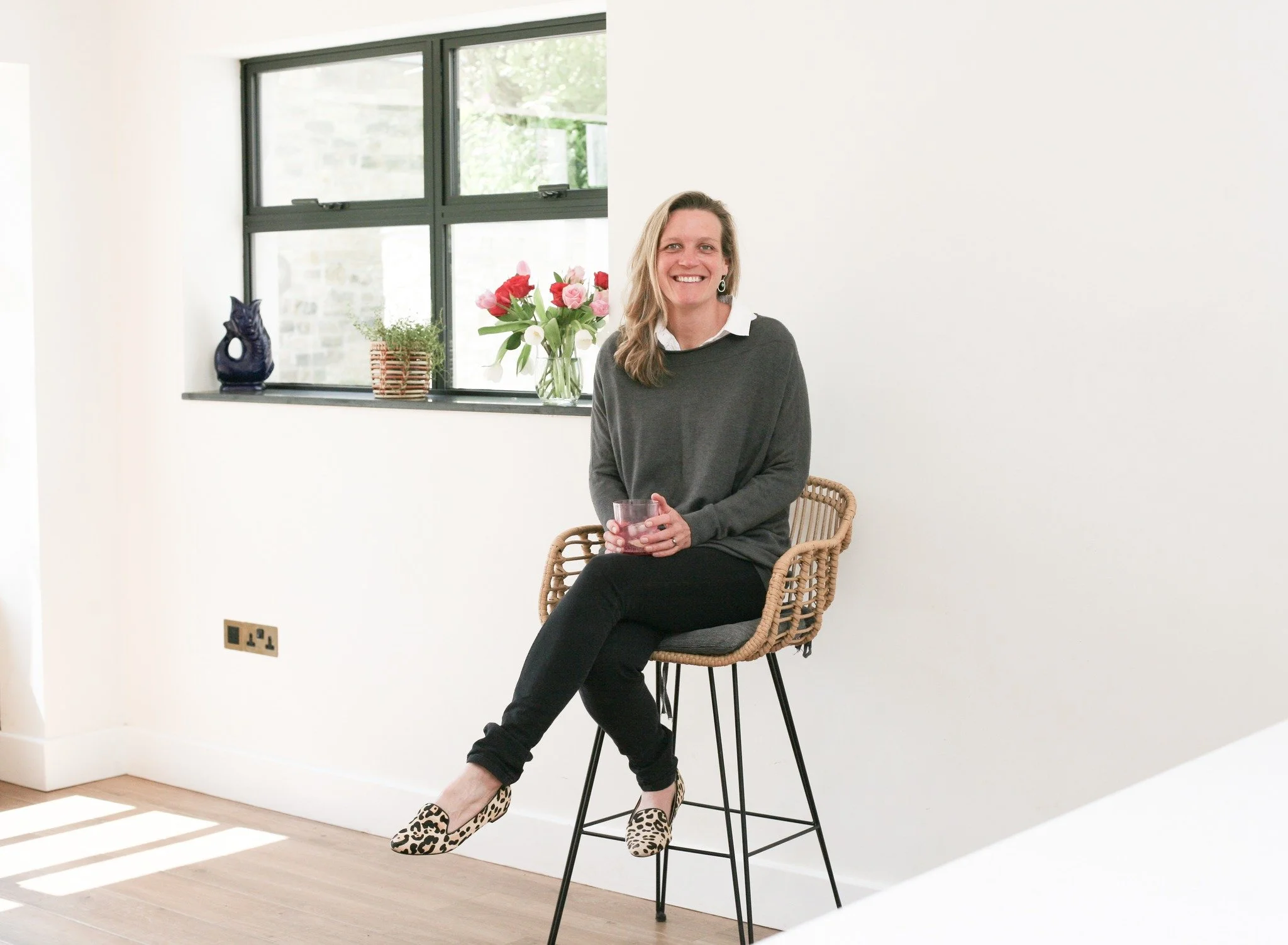 Annabel Gonifas sitting on a wicker chair indoors, smiling, holding a glass, wearing a white shirt, gray sweater, black pants, and leopard print shoes, with a window behind her displaying a vase of pink and white tulips and green plants.
