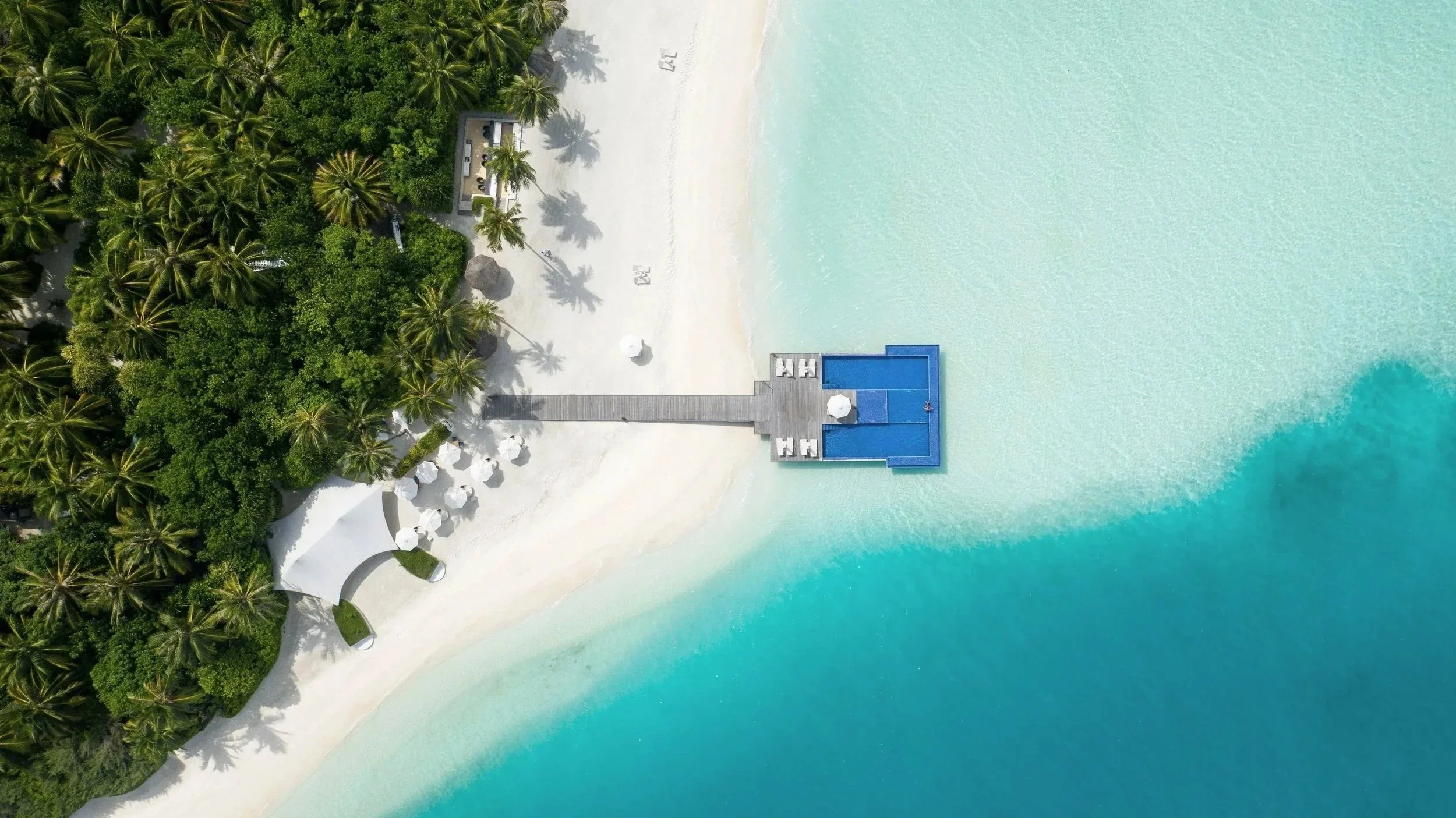 Aerial view of a tropical beach with white sand, lush green palm trees, and a blue ocean. There is a dock extending into the water with a structure at the end, and beach umbrellas and chairs on the sand.