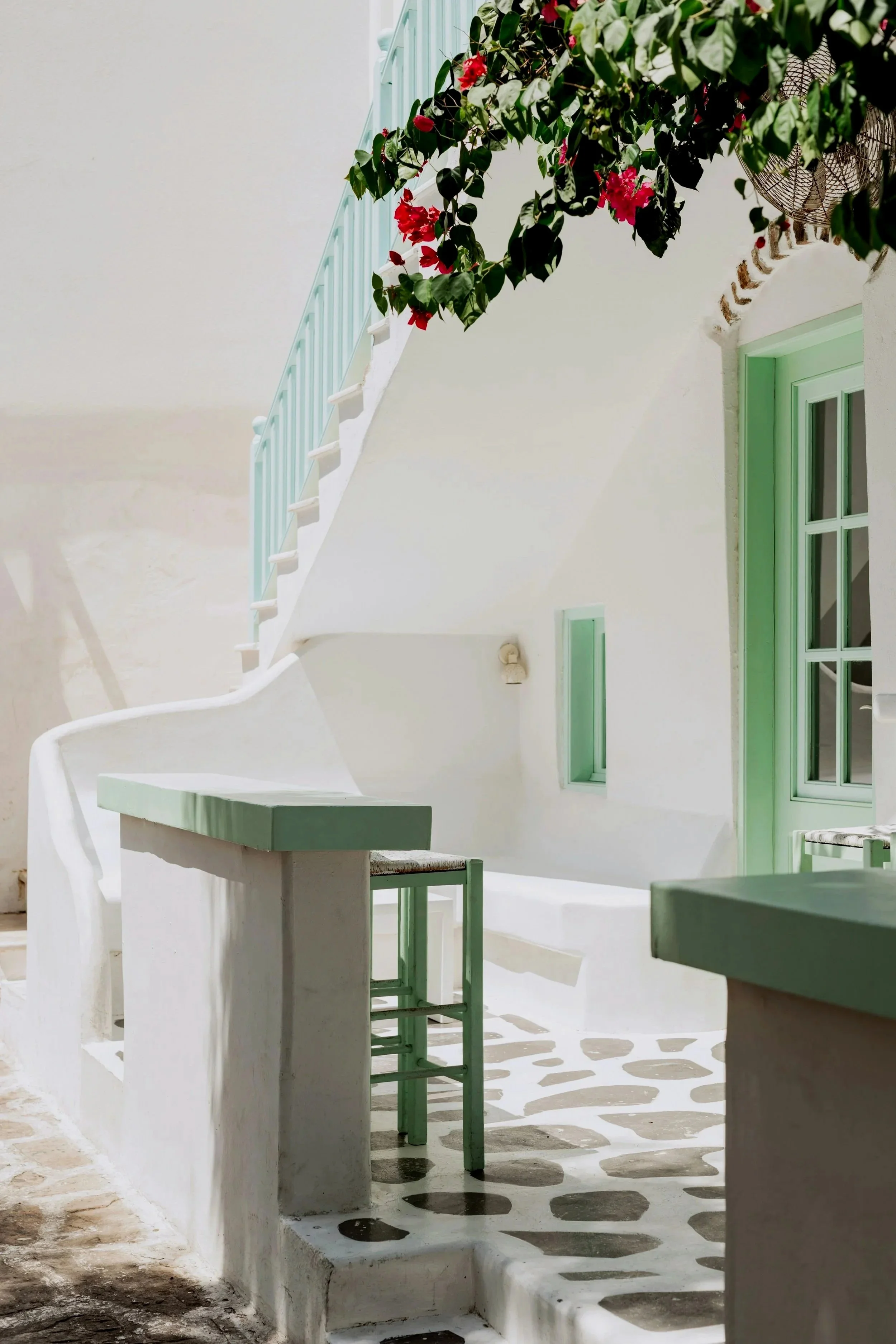 A white building with green window frames, a small staircase, and outdoor seating with a green chair. There are bushes with pink flowers and a fan visible. The ground has a patterned shadow.