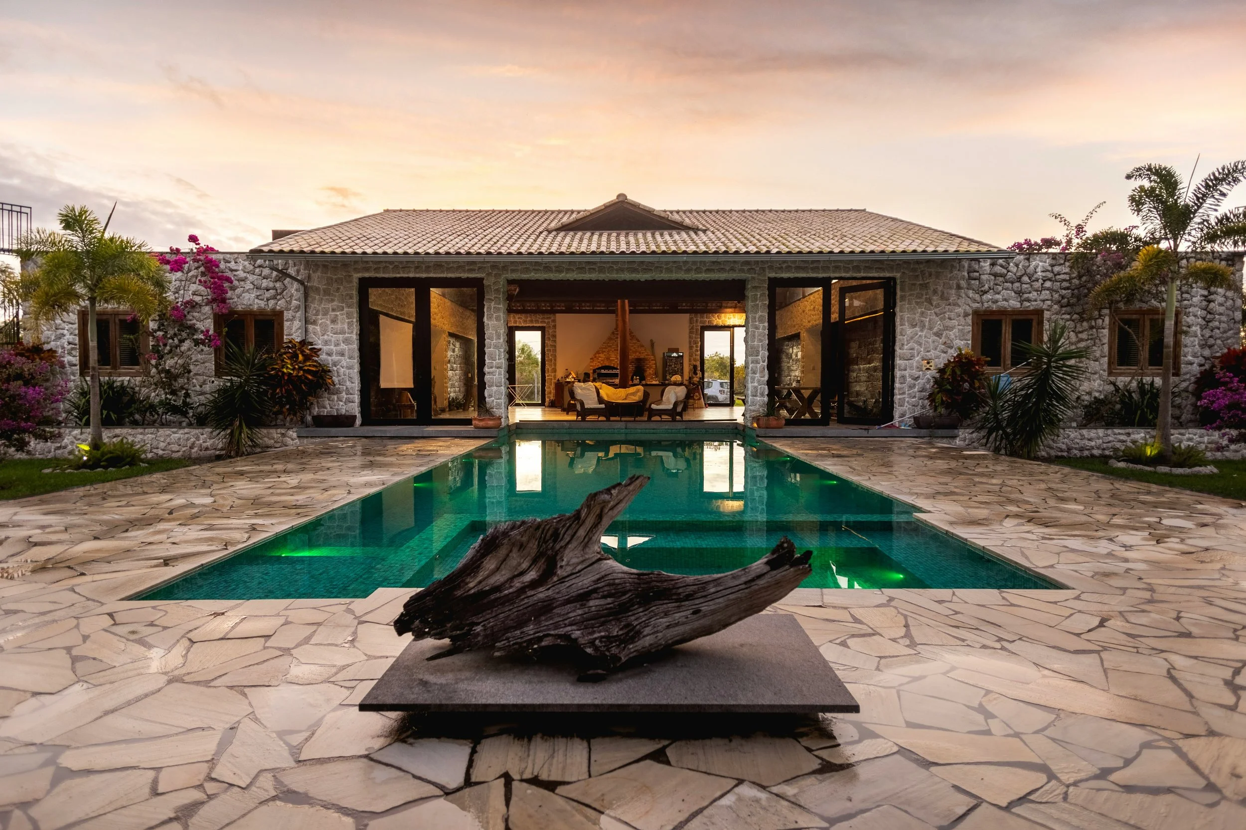 A stone house with a tiled roof and large glass doors opening to a patio with a swimming pool. There are palm trees and flowering plants around the pool area, with a sunset sky in the background.