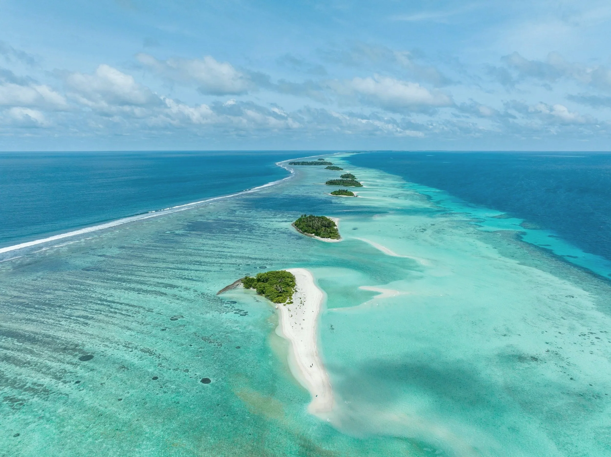 Aerial view of a chain of small islands and sandbars surrounded by turquoise ocean waters under a partly cloudy sky.
