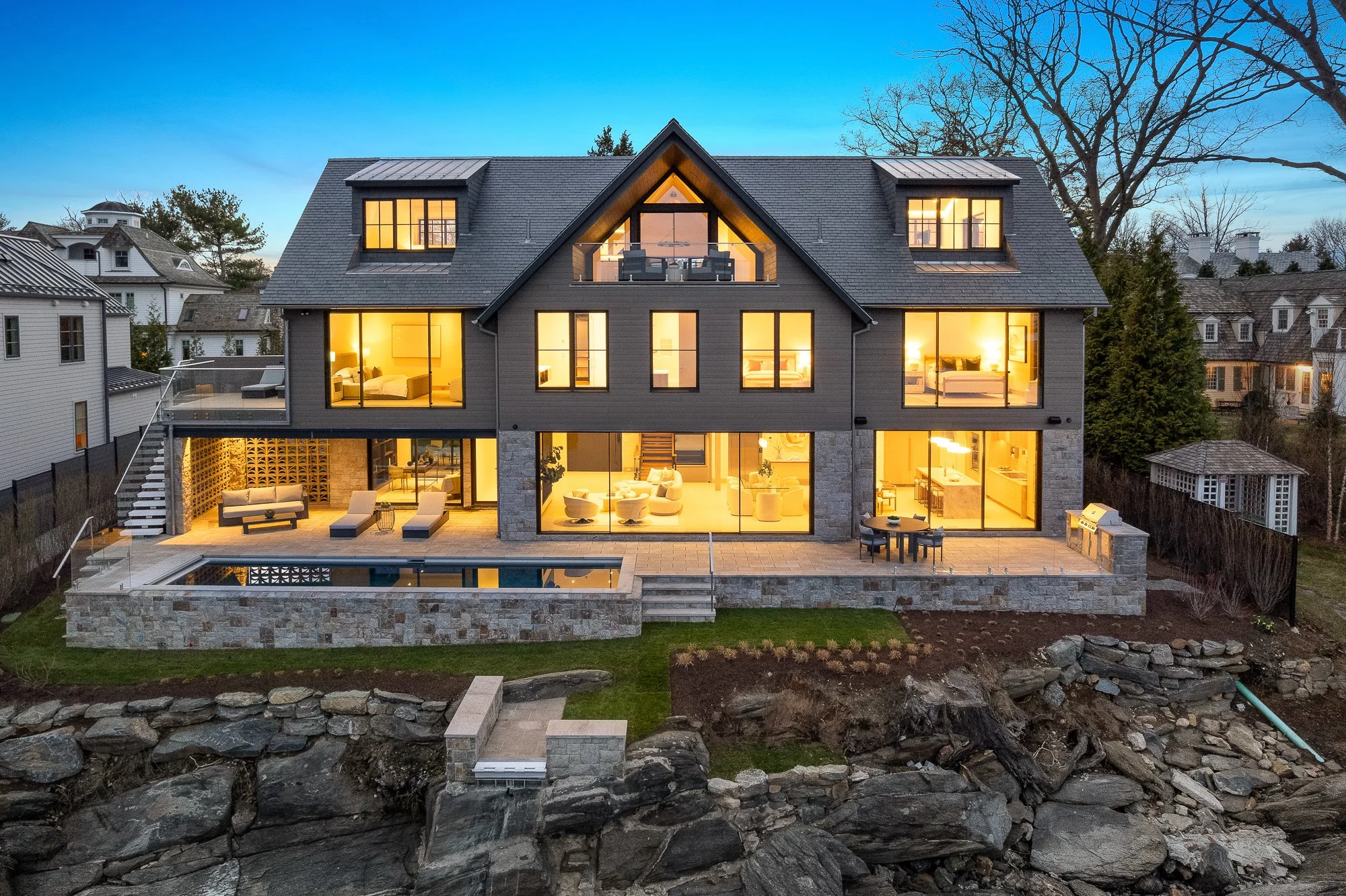 A modern multi-story house with large illuminated windows and a backyard patio with outdoor seating, overlooking a rocky shoreline in the evening.