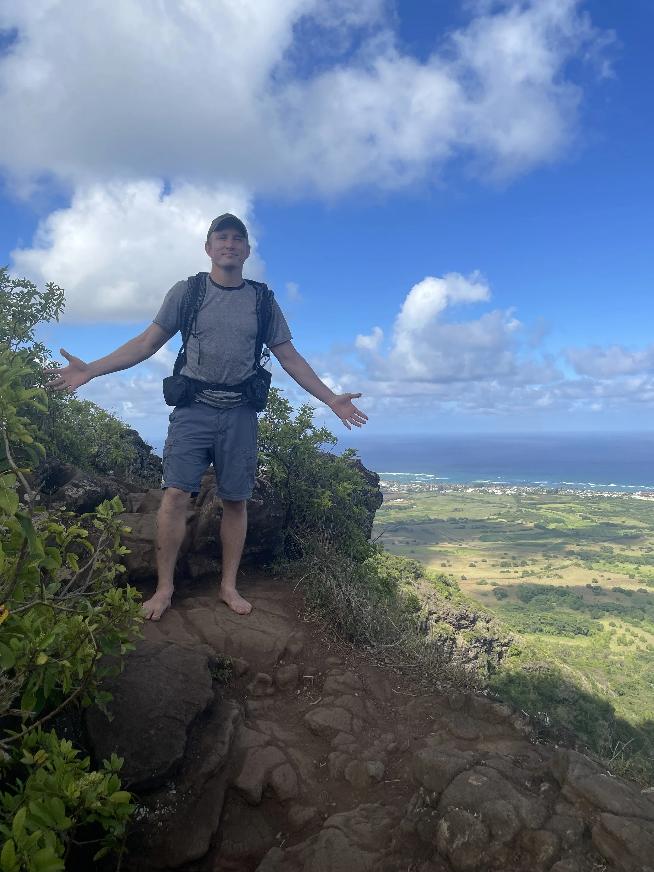 Will Ratliff standing on a rocky trail with open arms, overlooking a scenic view of green fields, a blue ocean, and partly cloudy sky.