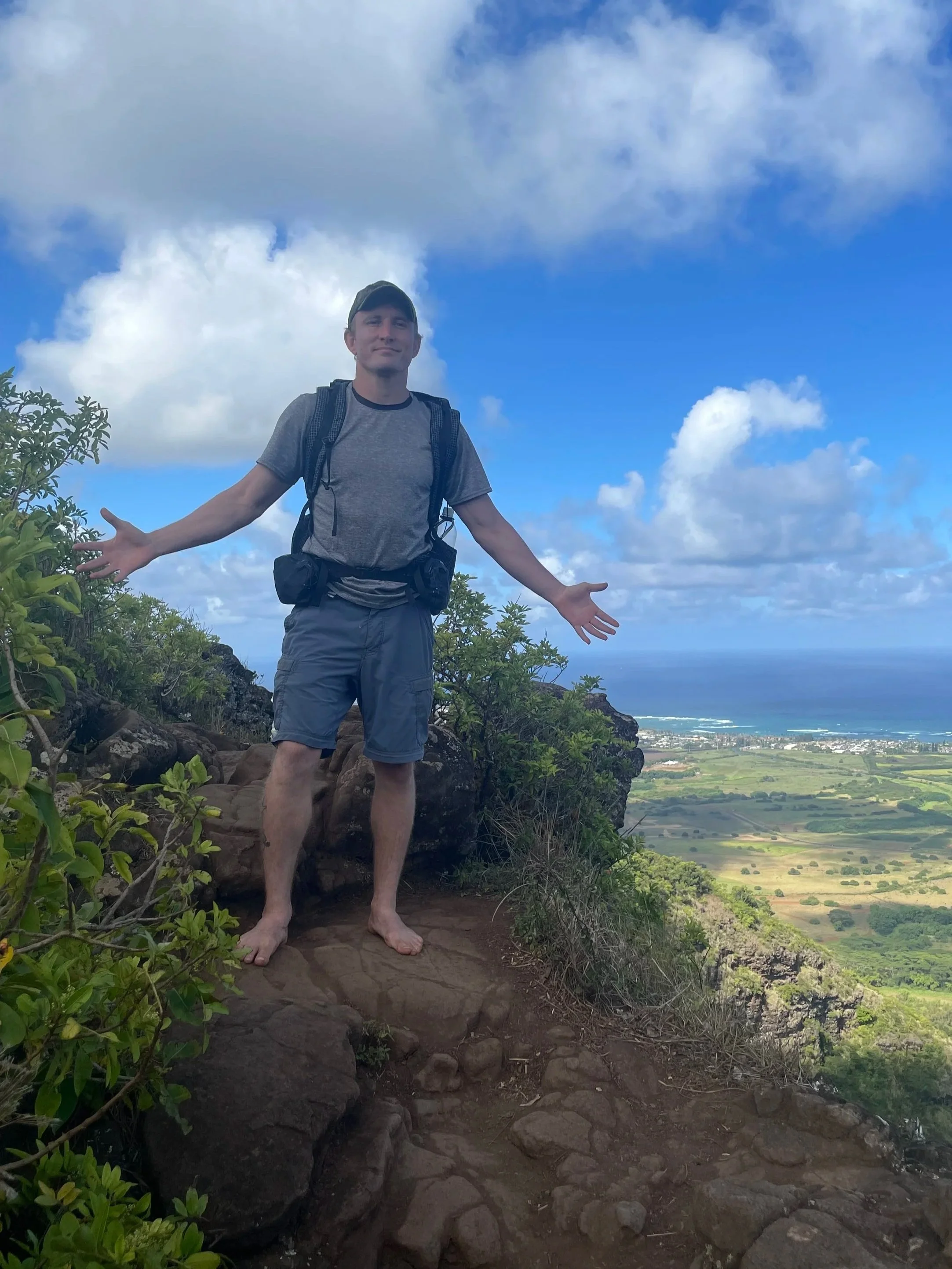 Will Ratliff standing on a rocky trail on a hilltop, stretching his arms out. He is dressed in gray shorts, a gray t-shirt, and a cap, with a backpack. The background shows a vast landscape with fields, a coastal line, and a partly cloudy sky.
