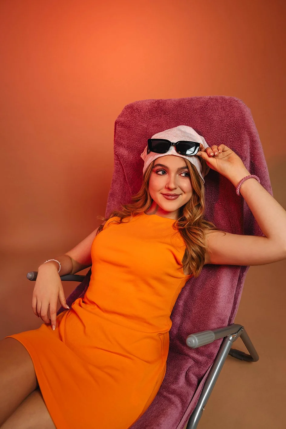 Stella Maria - Junge Frau in orange Kleid liegt entspannt auf einem Liegestuhl, trägt eine rosa Mütze, hält eine Sonnenbrille, und hat ein Lächeln auf dem Gesicht. Die Hintergrundfarbe ist warm.