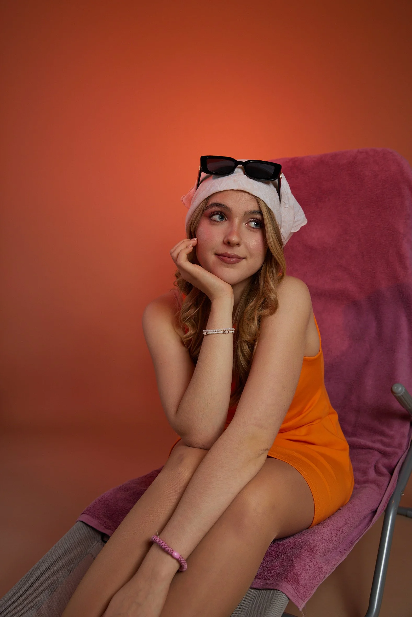 Junge Frau sitzt auf einem Stuhl mit lila Bezug, trägt einen weißen Schlaf- oder Schlafmütze, eine Suncara auf dem Kopf, ein orangefarbenes Oberteil und rosa Armbänder, vor einem orangefarbenen Hintergrund.