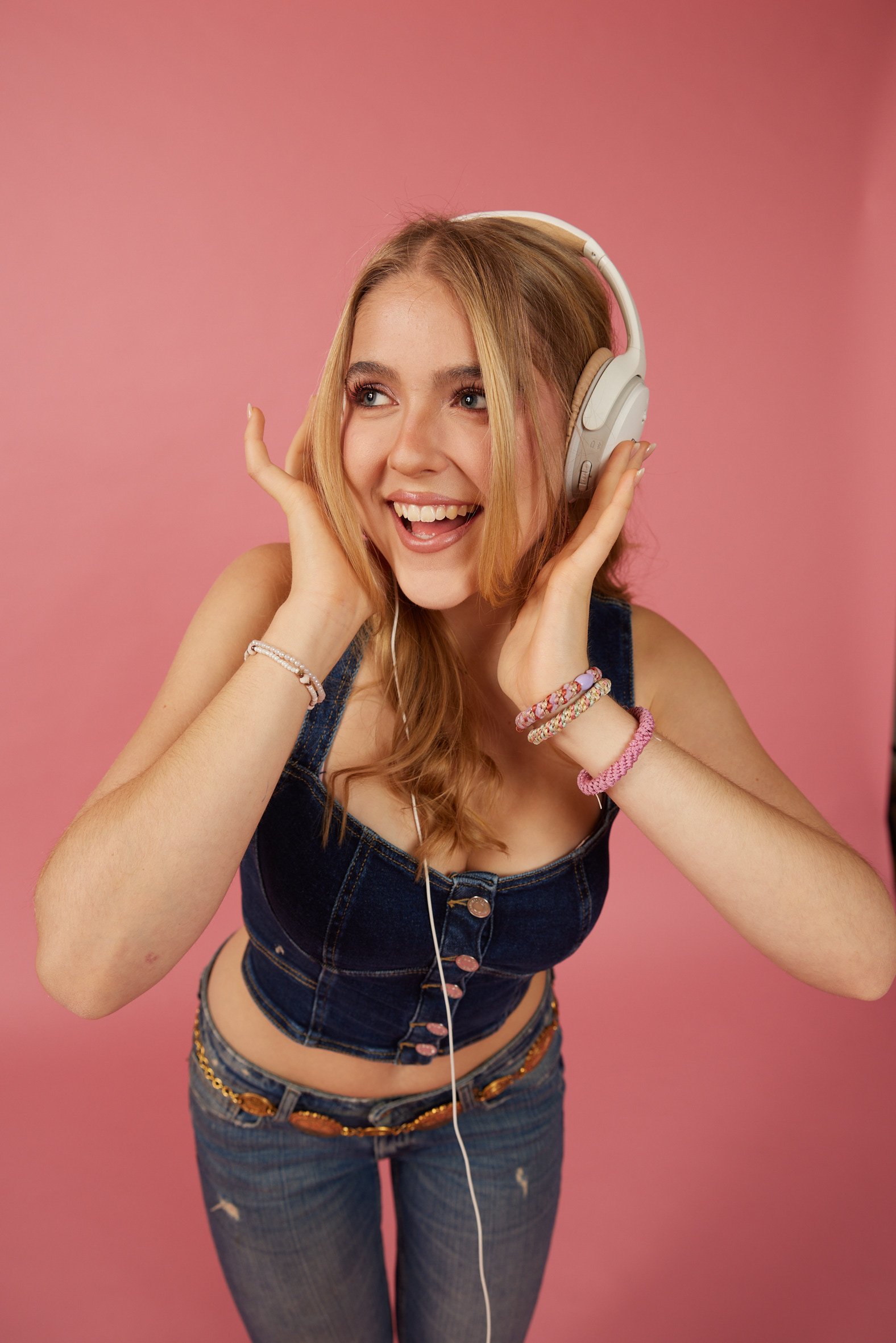 Stella Maria - Junge Frau mit roten Haaren trägt Kopfhörer, lächelt und tanzt vor rosa Hintergrund, trägt Jeans und ein Top, accessorized mit Armbändern und Ketten.