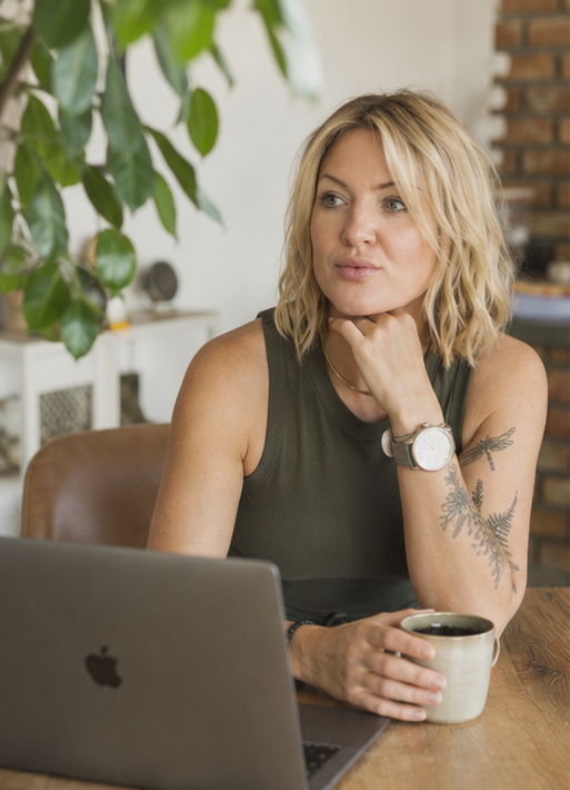 A woman with blonde wavy hair sitting at a wooden table with a laptop and holding a mug, in a cozy room with green indoor plants and a brick wall in the background.