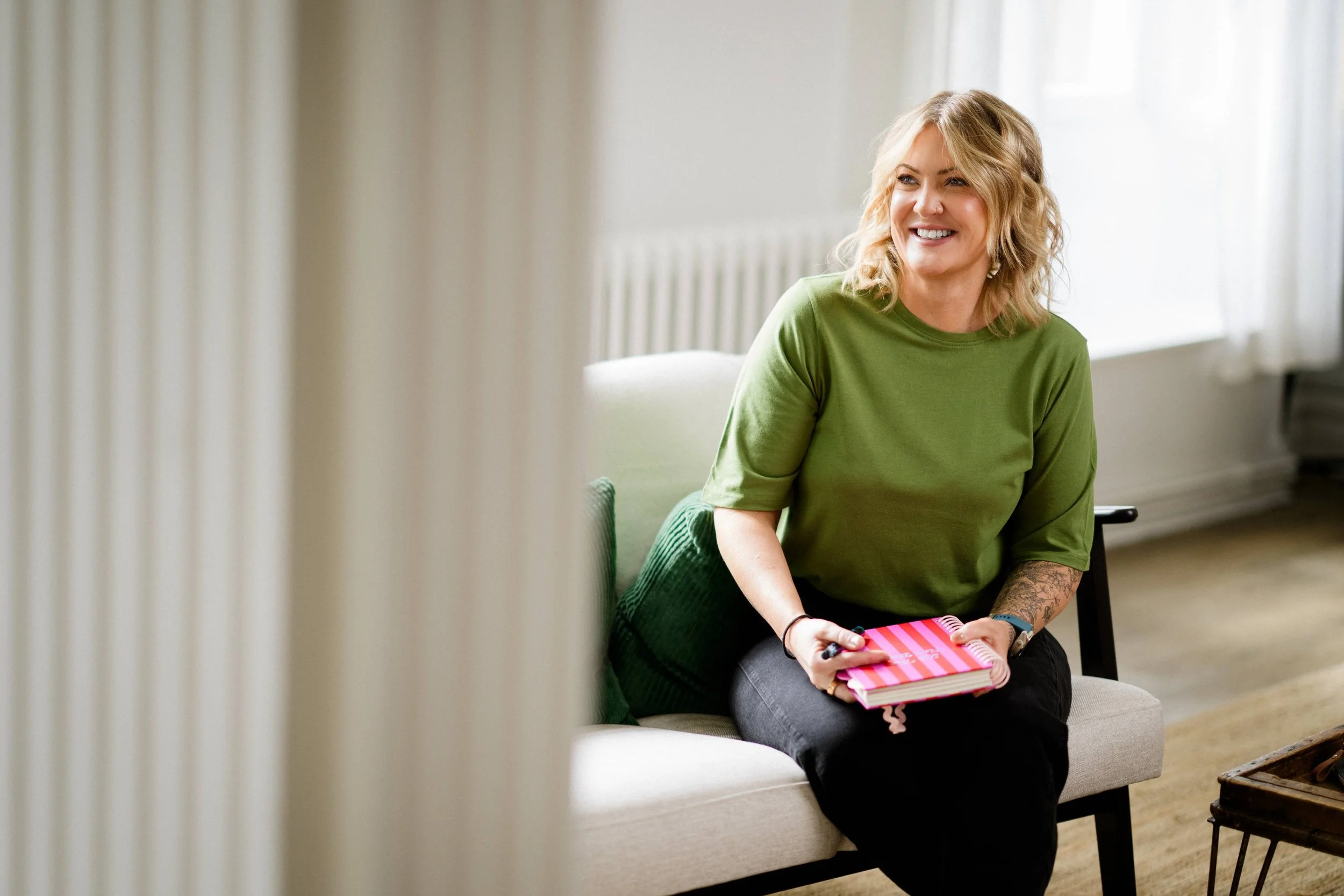 A woman with blonde hair and an olive green sweater speaking to a group of seated diverse women in a bright, modern room with large windows and plants.