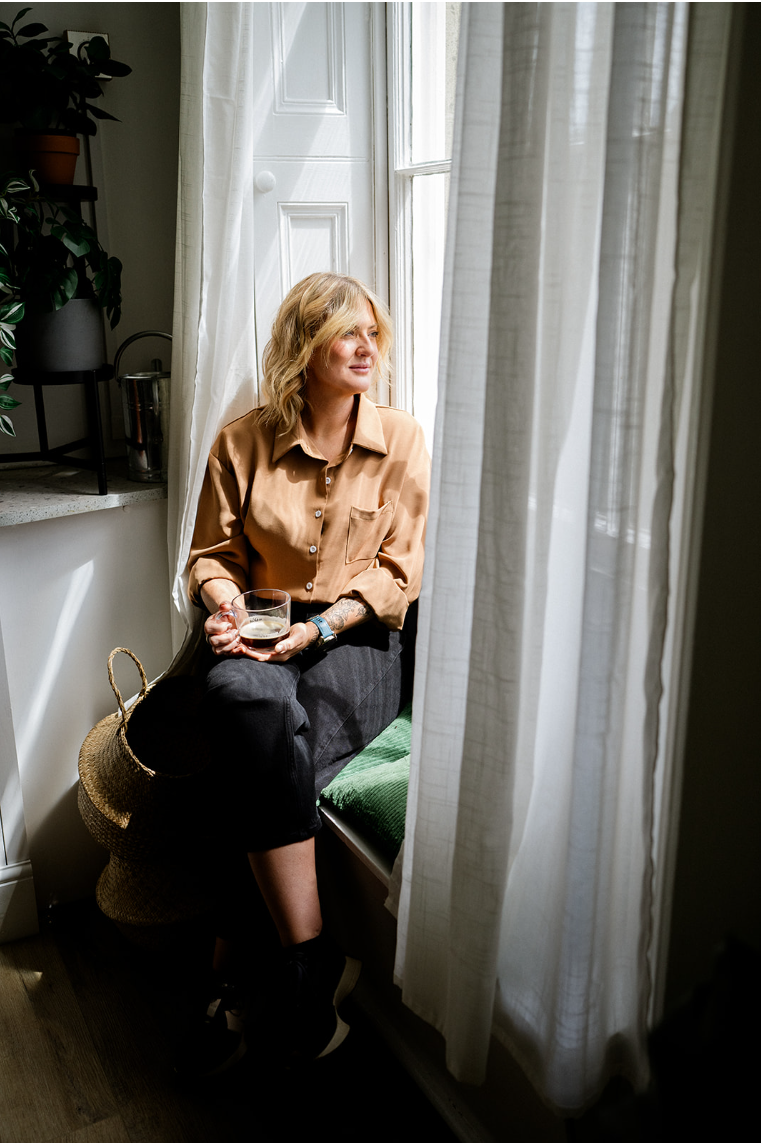 A woman with blonde, wavy hair sits on a window seat, holding a glass of water, gazing outside with a peaceful expression. She wears a light brown button-up shirt and dark pants, with a straw bag hanging on her side. The room is bright, with sunlight coming through sheer white curtains and a window beside her.