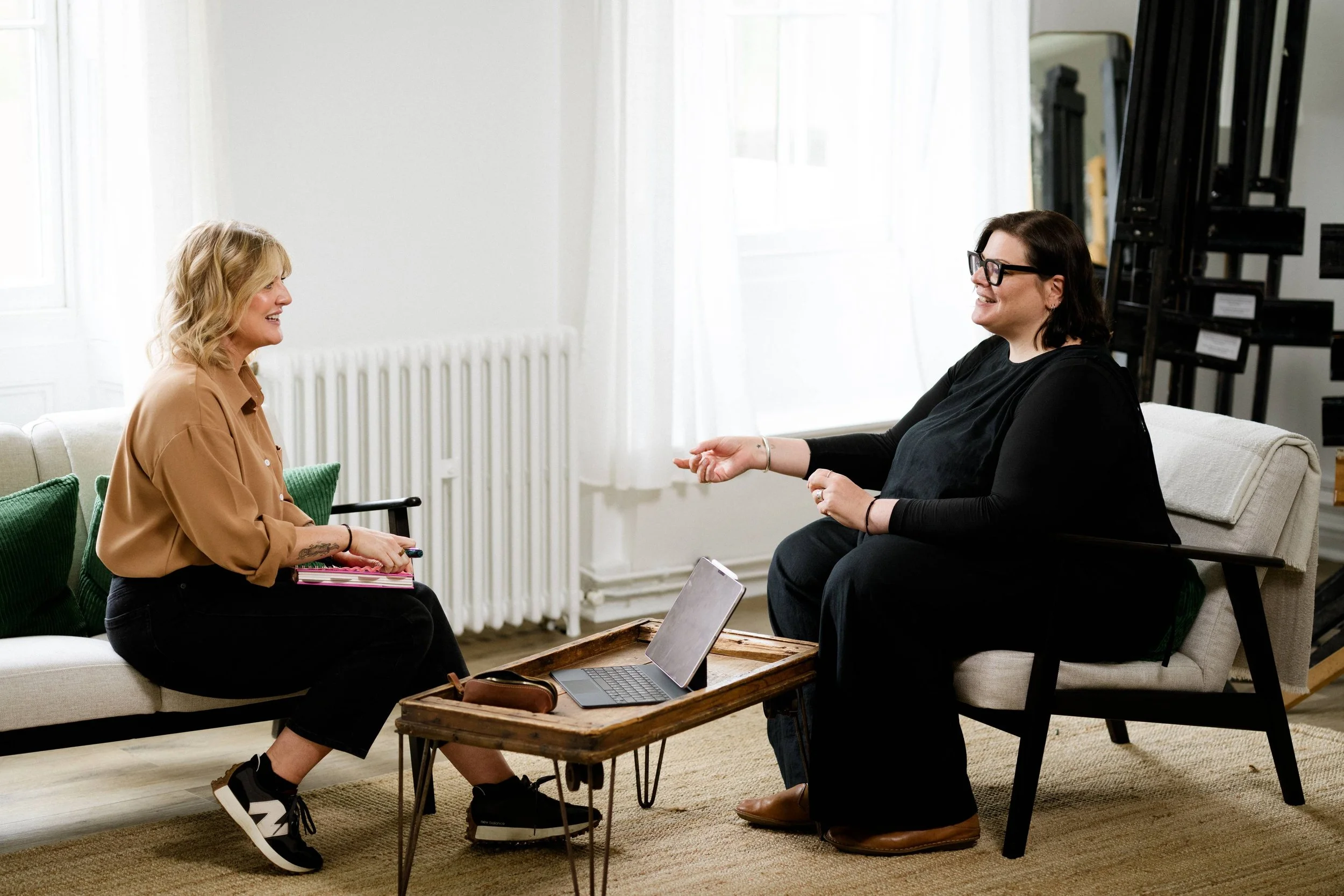 Two women having a conversation in a living room. One woman is sitting on a sofa, holding a notebook, and smiling. The other woman is sitting on a chair, wearing glasses, and gesturing with her hand. There is a laptop on a wooden tray table between them.