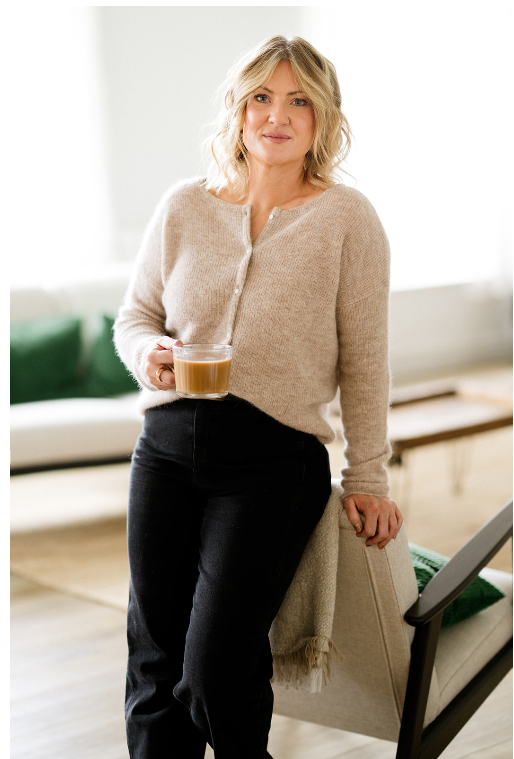 A woman stands in a bright living room, holding a glass of coffee, wearing a beige sweater and black pants.