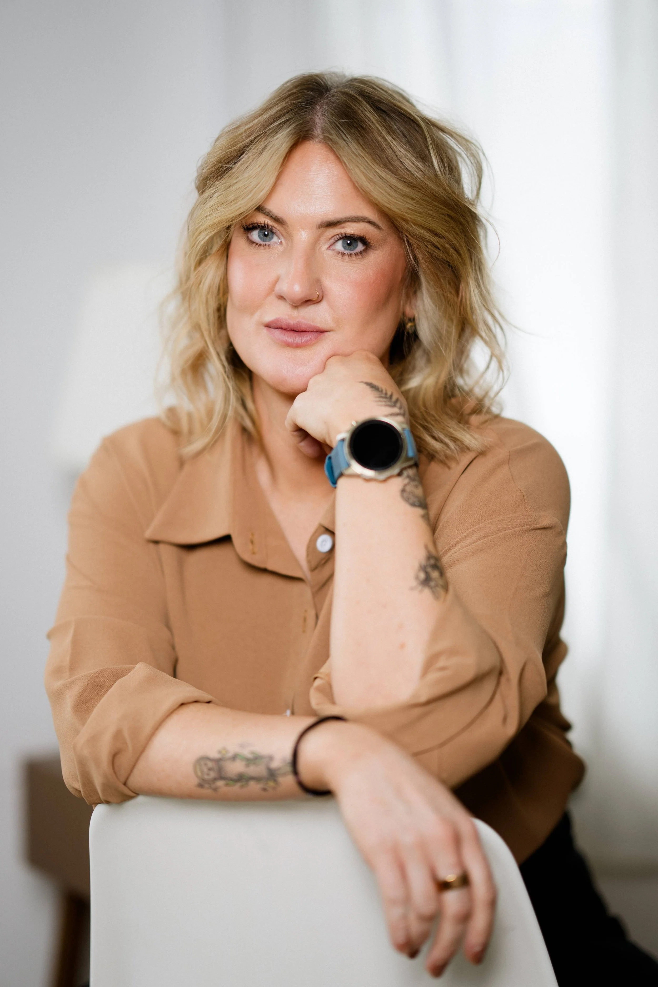 A woman with blonde, wavy hair and blue eyes, resting her chin on her hand, wearing a tan shirt, with tattoos on her left arm and a watch on her right wrist, sitting behind a white chair against a plain, light background.