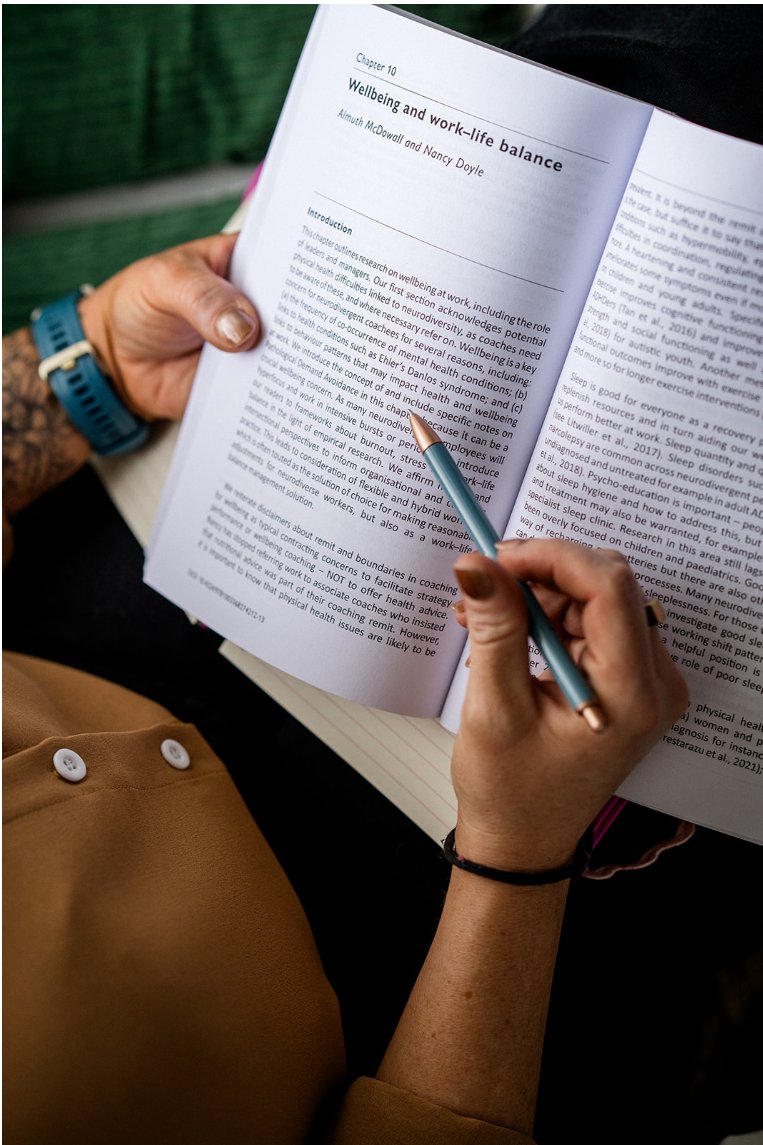 Person holding an open book, pointing to a chapter titled 'Wellbeing and work-life balance' with a blue pen, wearing a tan sleeve and a blue watch.