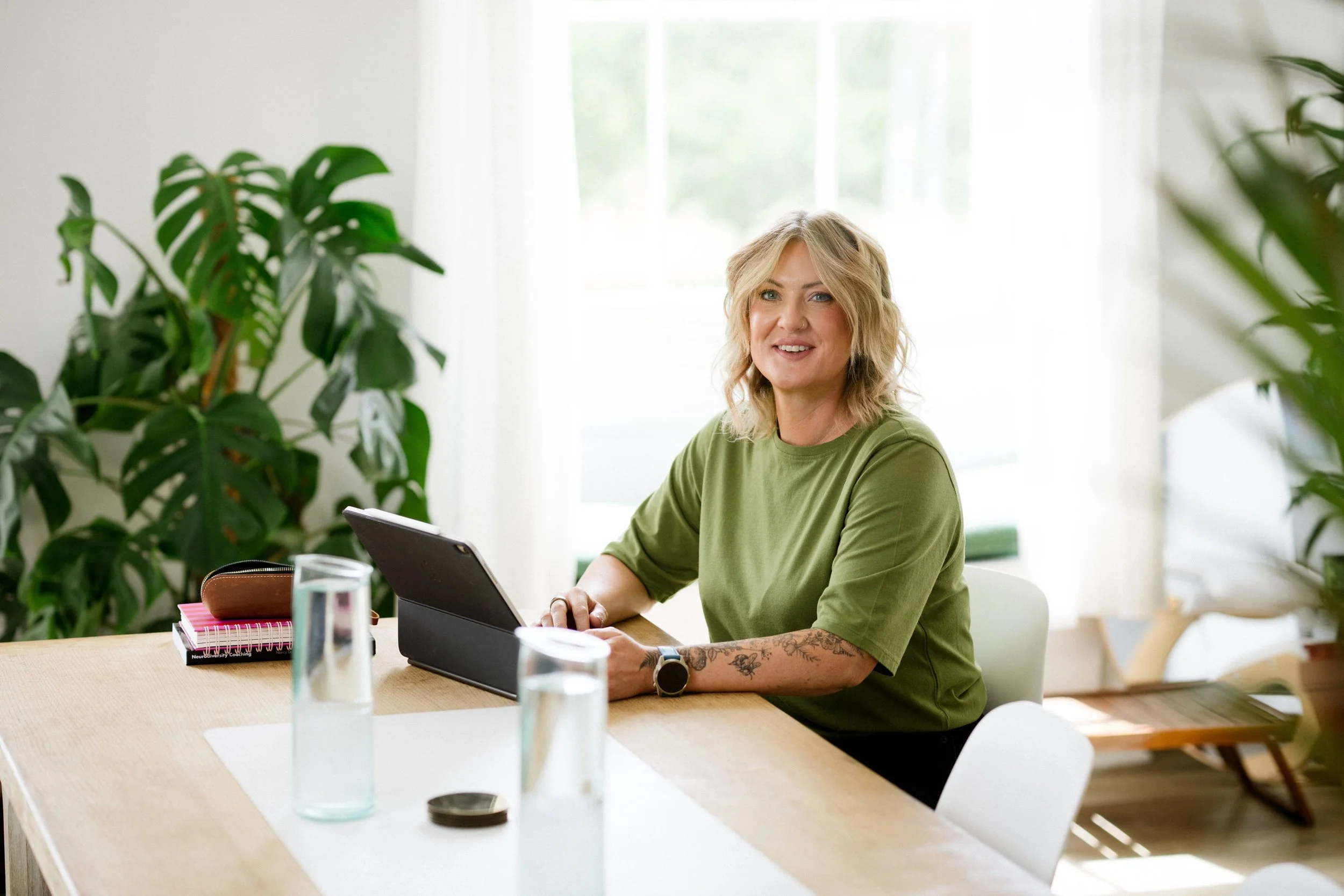 A woman with blonde hair and a nose piercing sitting at a table with a tablet, surrounded by green plants, in a bright room with a large window.
