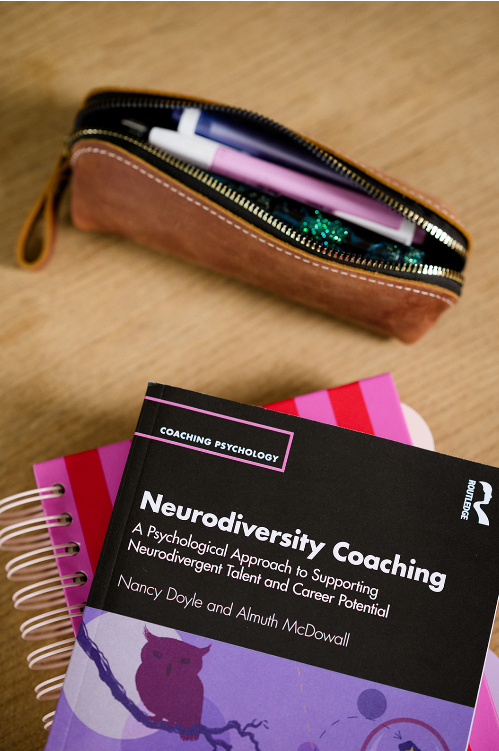 Open brown zipper pouch on wooden surface with pens and highlighters inside, next to a pink spiral notebook with a black book titled "Neurodiversity Coaching" in psychological coaching.