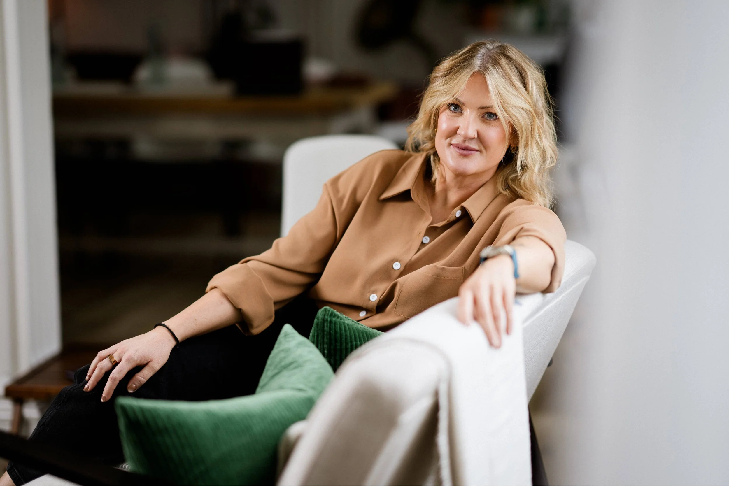 A woman with blonde hair tied in a loose ponytail, wearing a green sweater and earrings, sitting in a wooden chair with a smile on her face, indoors with a beige wall and a potted plant in the background.