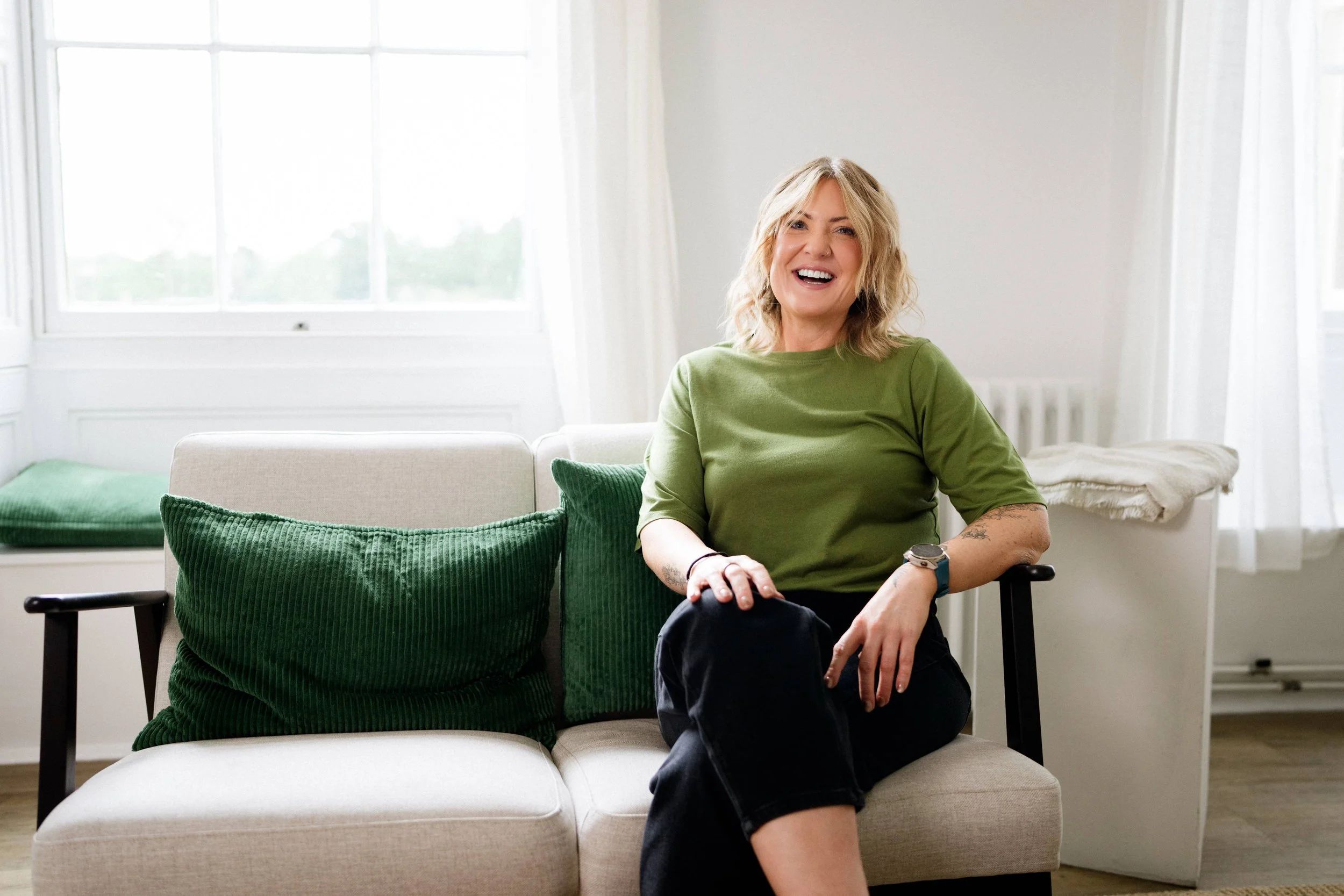 A smiling woman with blonde hair wearing a green shirt, sitting on a beige couch in a bright room with large windows and white curtains.