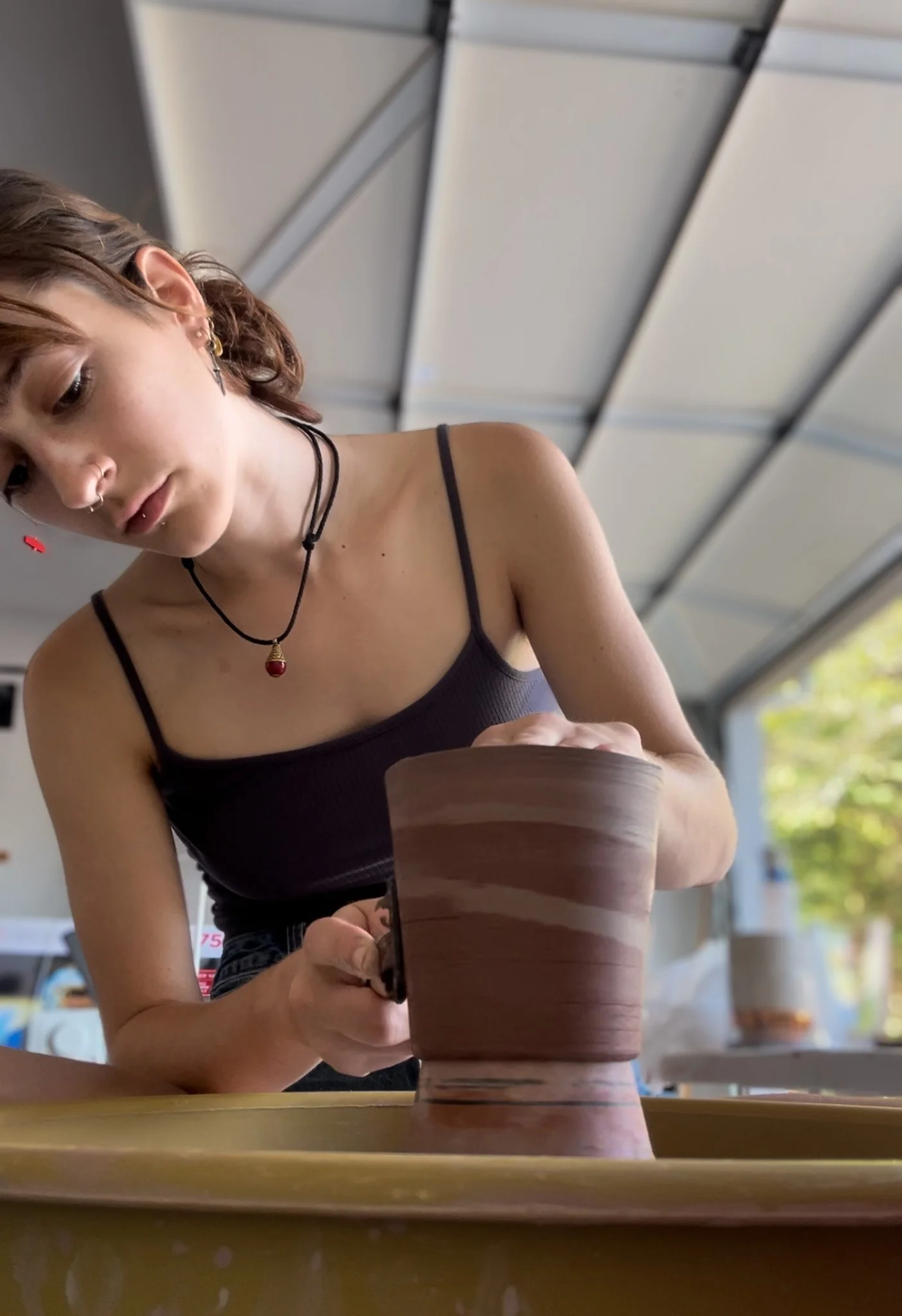 A young woman with short, wavy hair, wearing a black spaghetti strap top and jewelry, is sculpting a large clay pottery piece in a pottery studio. She is focused on her work, and there is pottery and tools around her. The background shows a modern industrial ceiling with beams and some windows.