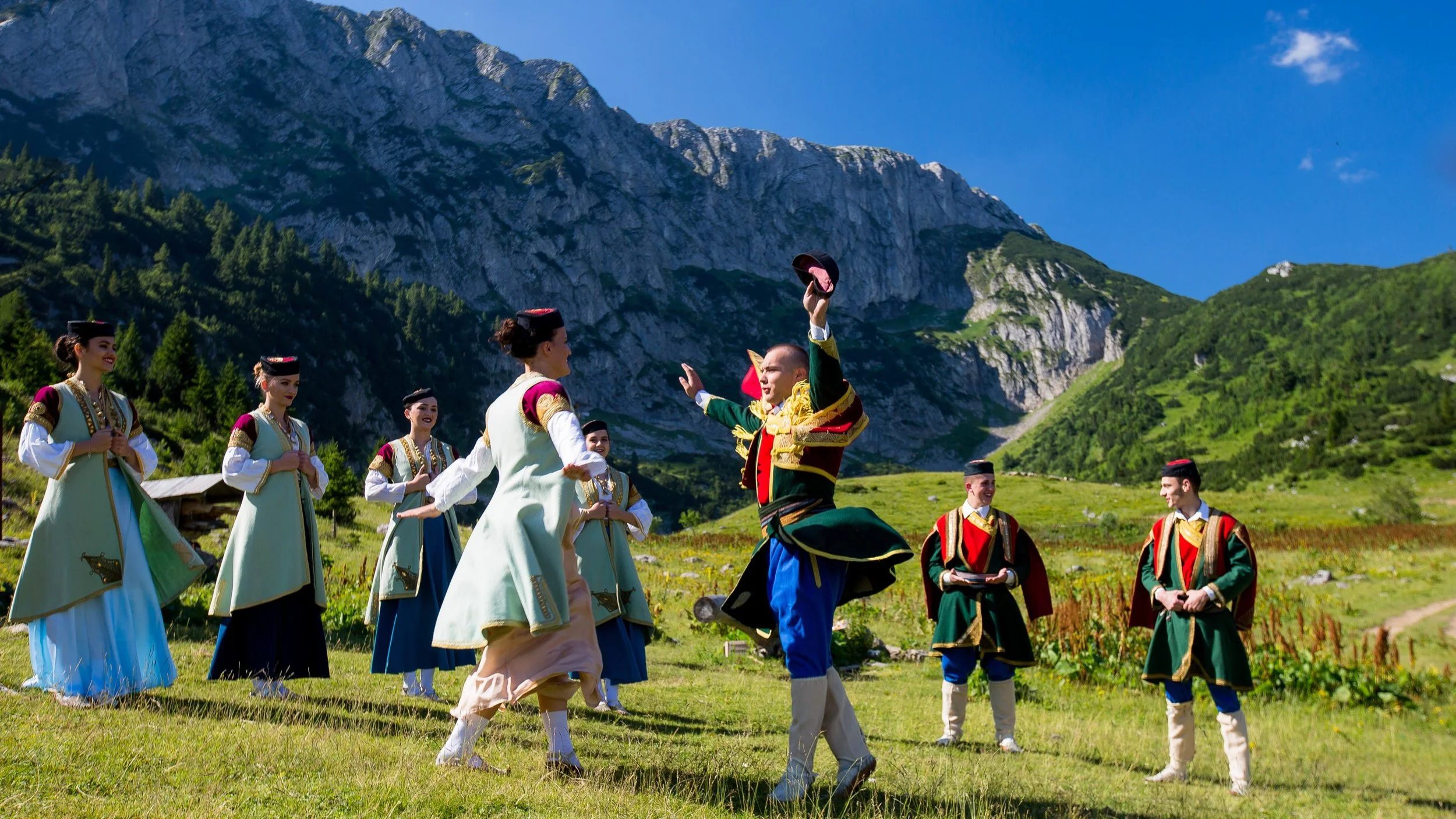 People in traditional clothing perform a dance outdoors in a scenic mountainous area with green hills and rocky peaks under a clear blue sky.