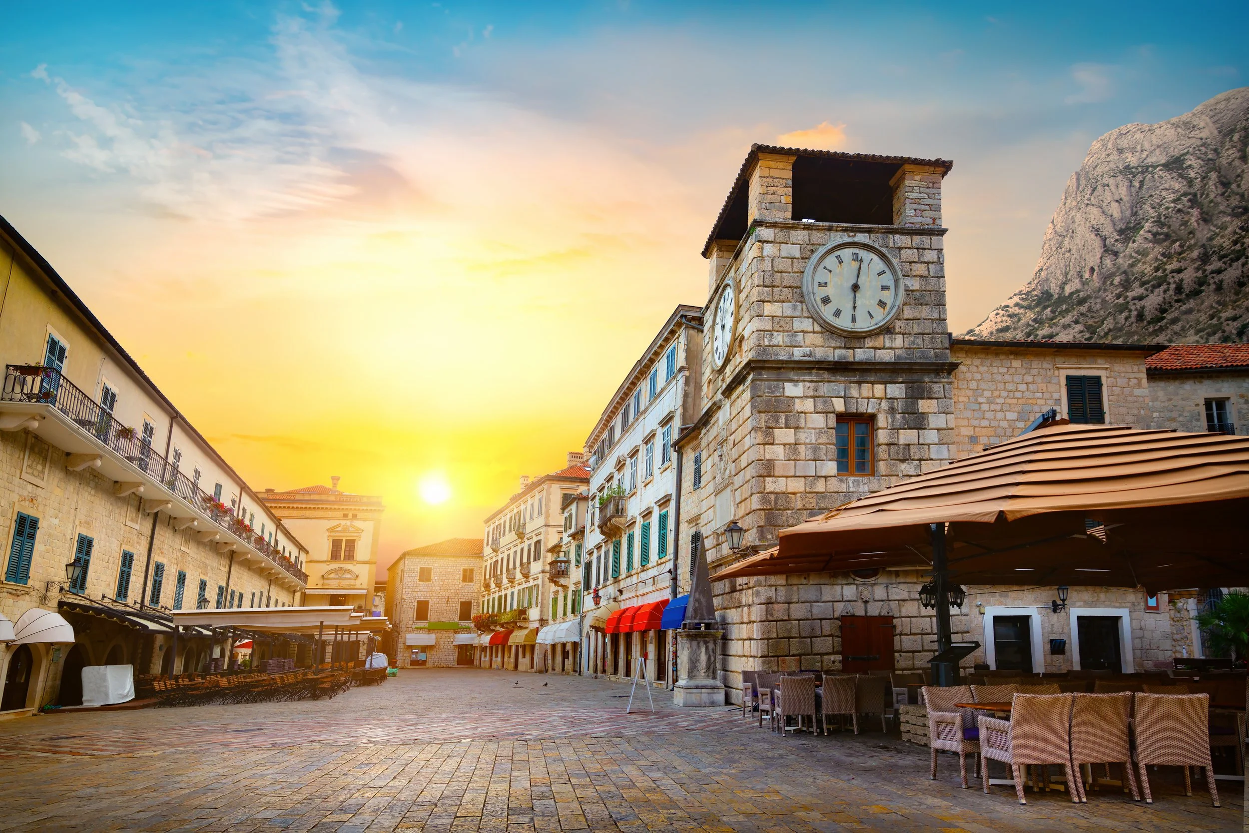 Sunset over a historic European town square with cobblestone streets, outdoor cafes, and a prominent stone clock tower, surrounded by old buildings with colorful awnings and a mountainous backdrop.