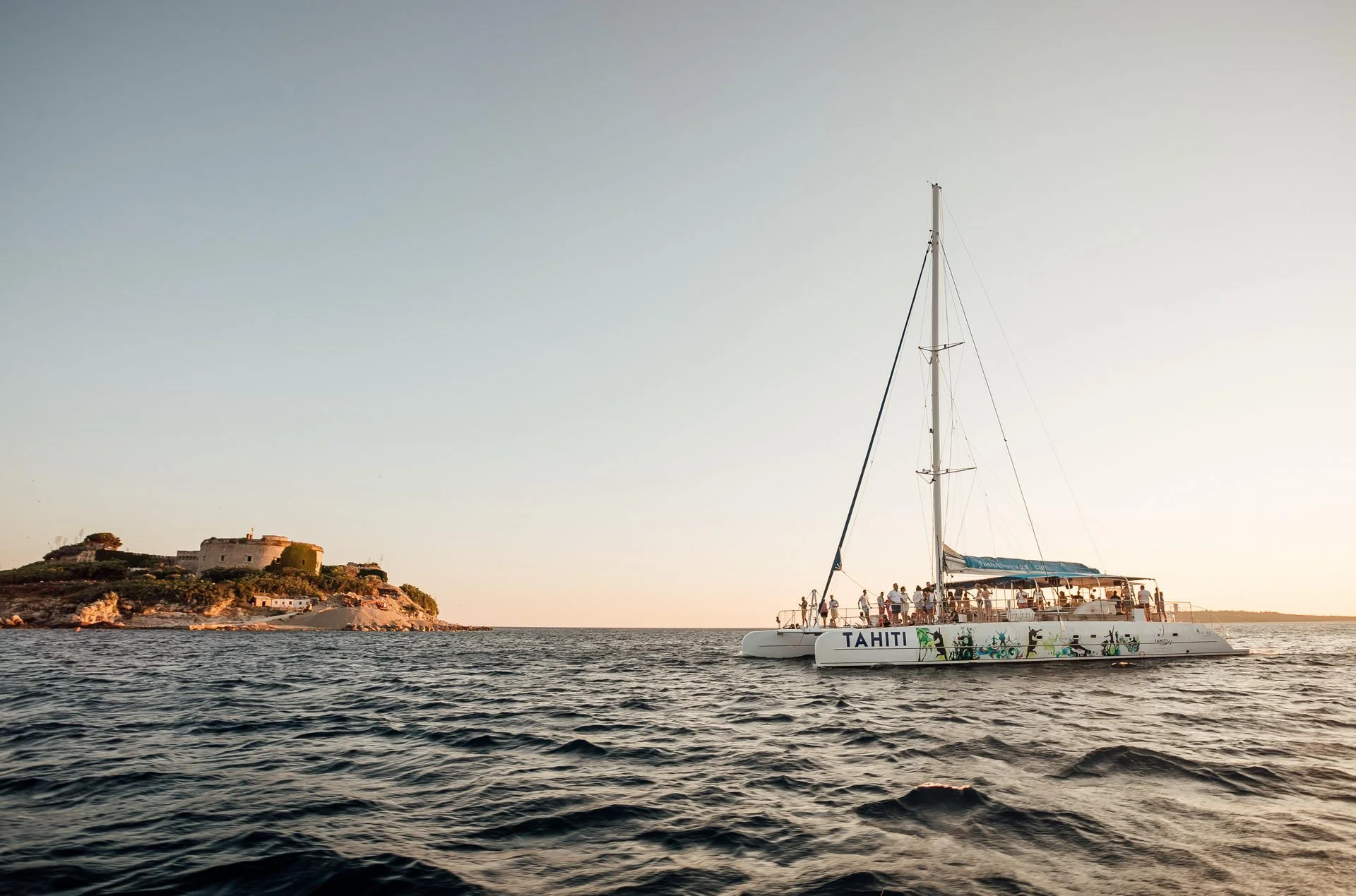 A white sailing catamaran with colorful artwork and the word 'Tahiti' written on its side, floating on calm water during sunset with people onboard and old buildings on a hill in the background.