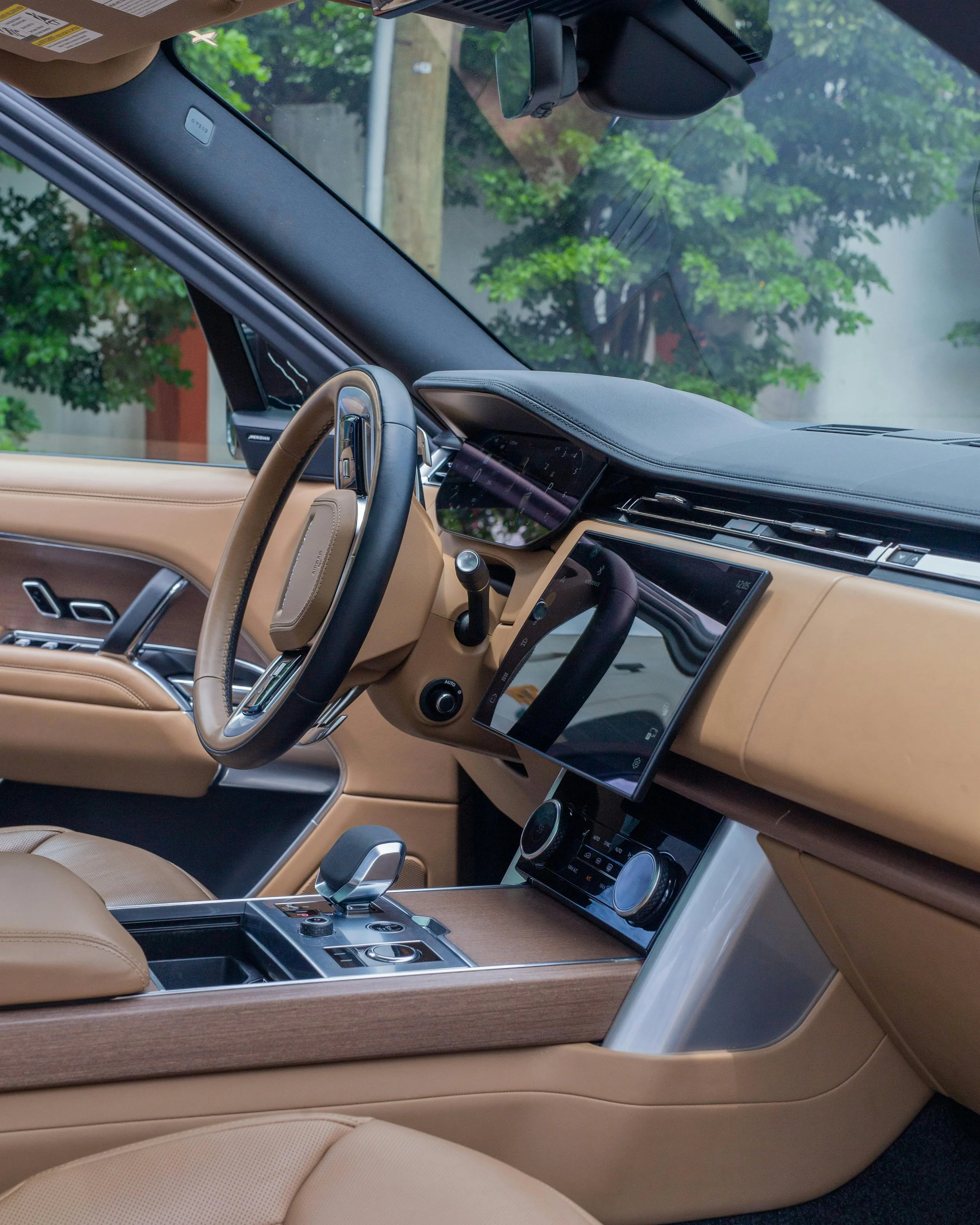 Interior of a luxury car with beige leather seats, a digital dashboard, and a modern central console with controls, seen from the driver's side. There is a large touchscreen display and a steering wheel with metallic accents. The windshield shows greenery outside.