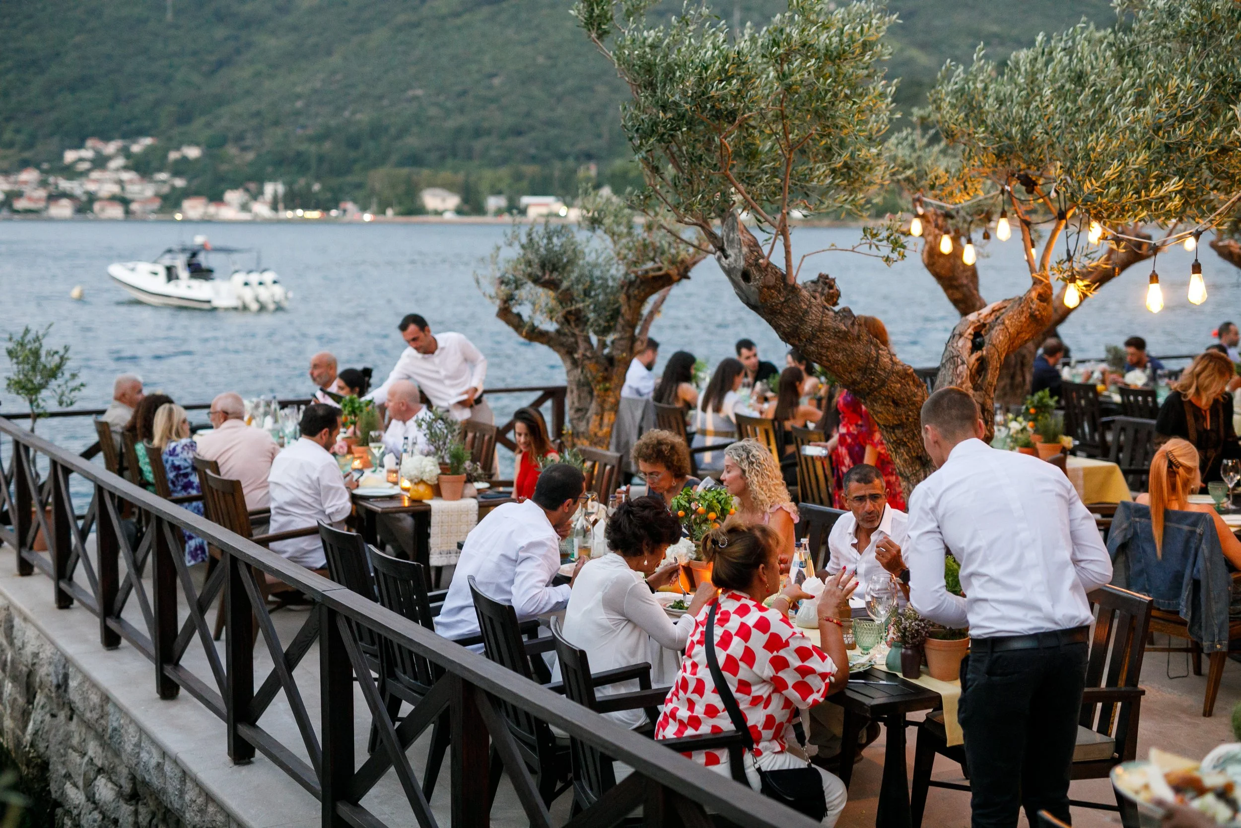 An outdoor gathering of people dining at tables by a lakeside during the evening, with hills and houses across the water, illuminated trees, and a boat on the lake.
